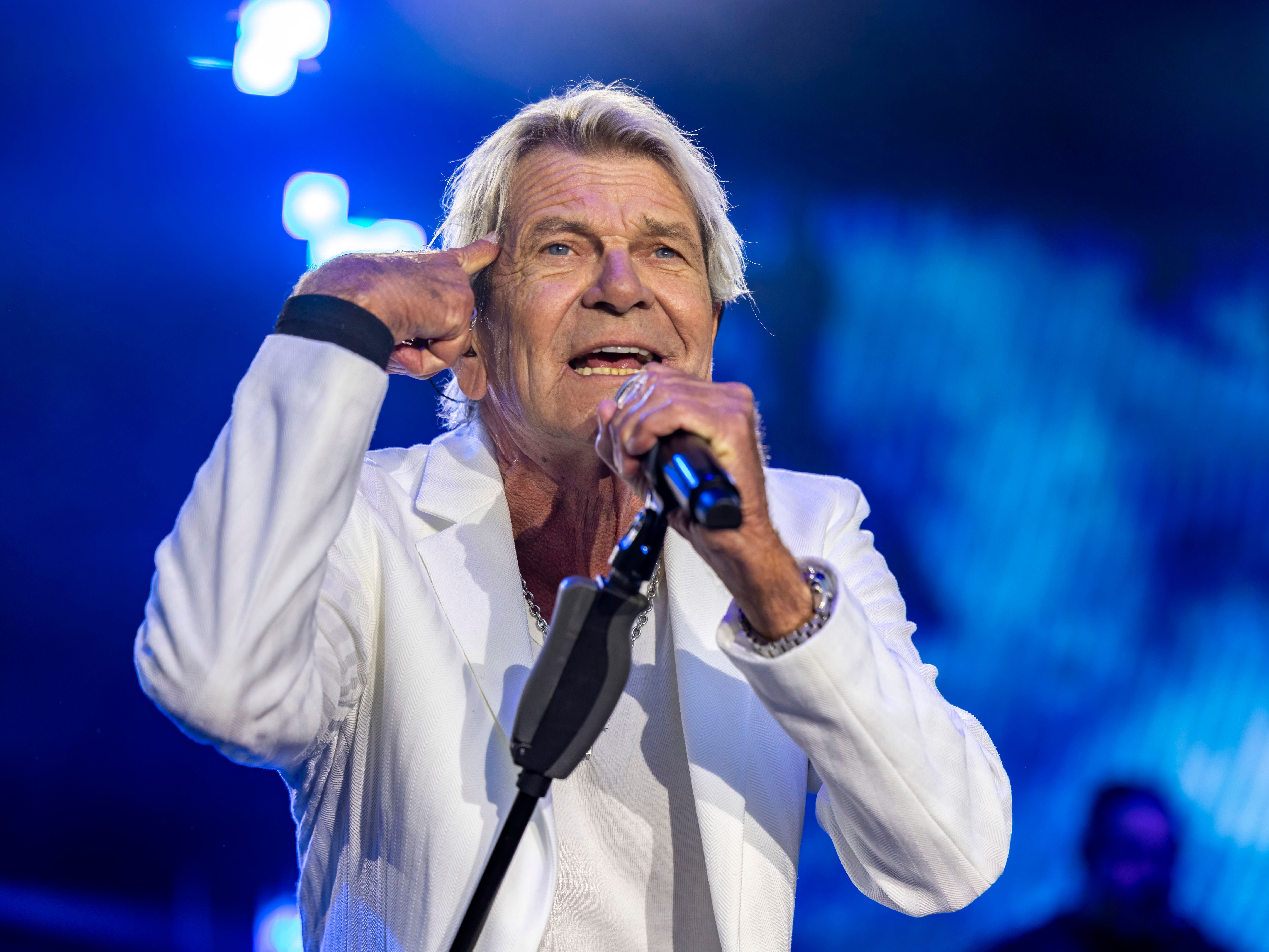 Ein älterer Mann im weißen Anzug singt live auf einer Bühne mit blauem Licht im Hintergrund.