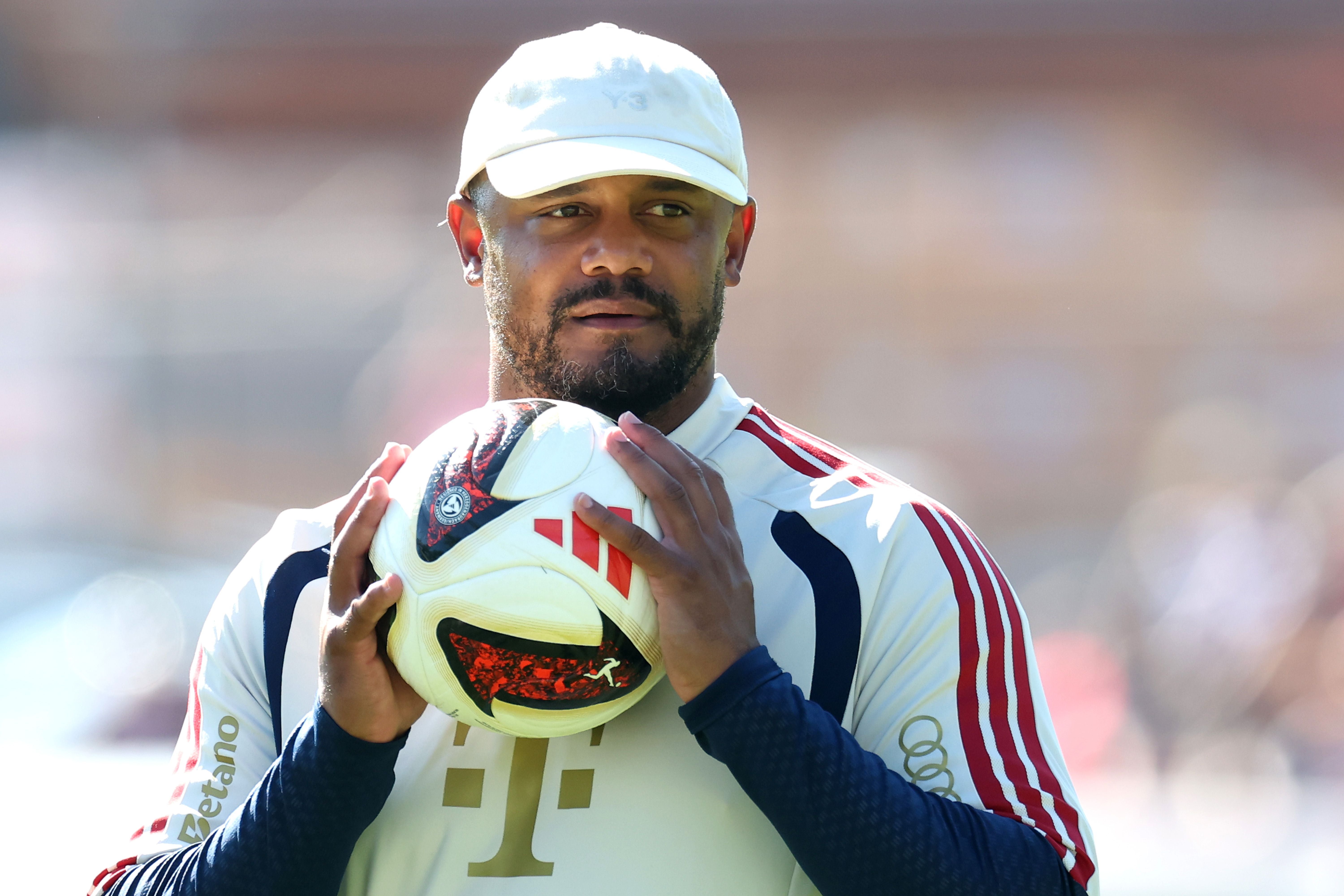 Ein Mann im Trainingsanzug hält einen Fußball und trägt eine weiße Kappe.