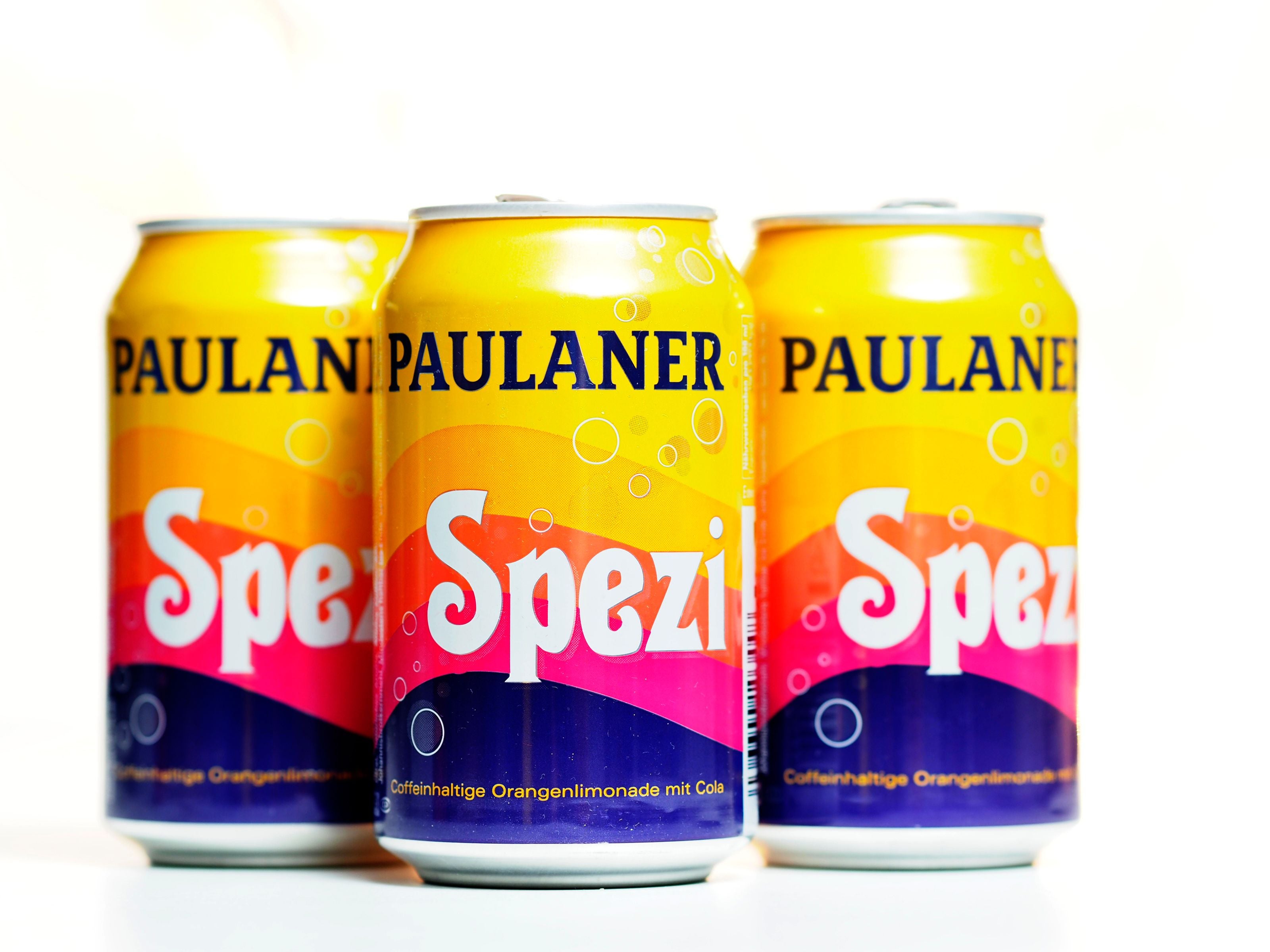 Drei Dosen Paulaner Spezi stehen nebeneinander auf weißem Hintergrund.