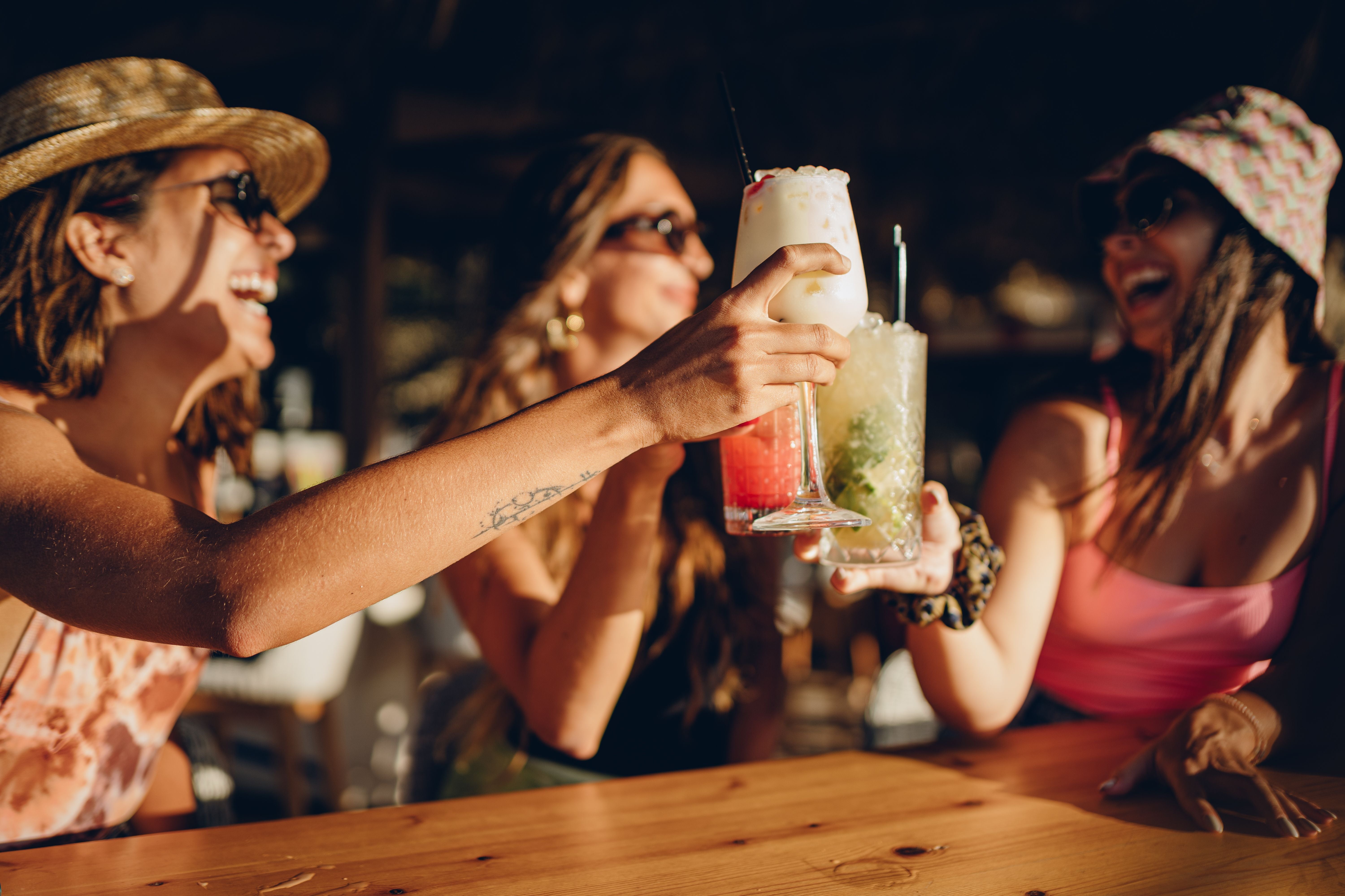 Drei Frauen stoßen fröhlich mit Cocktails an einem Holztisch an.