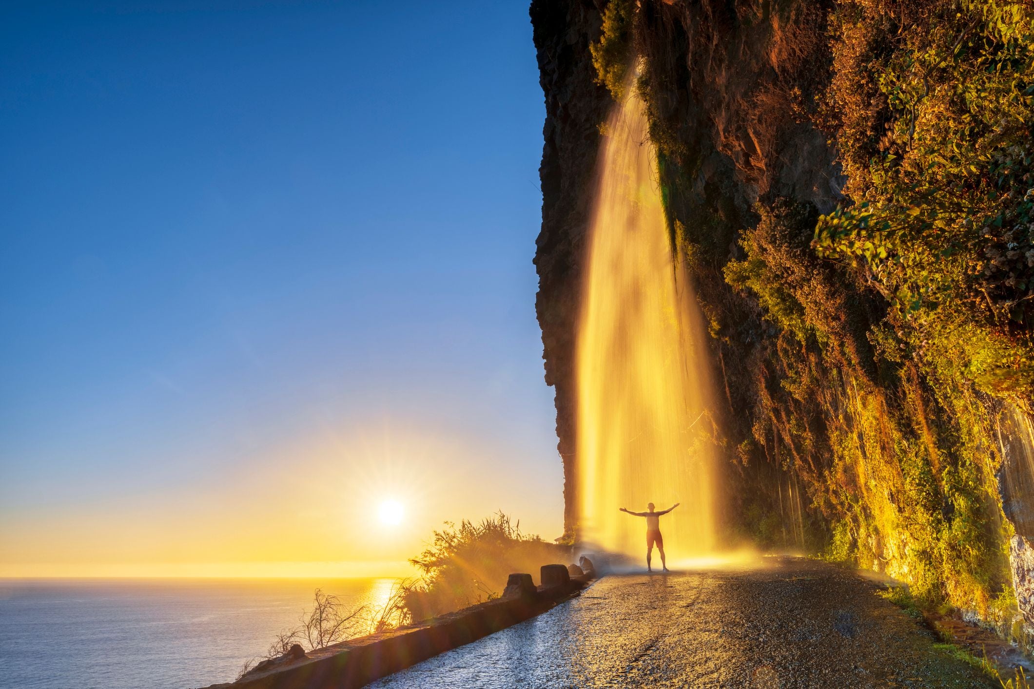 Mann steht beim Sonnenuntergang unter einem Wasserfall an einer Klippe auf Madeira, Portugal.