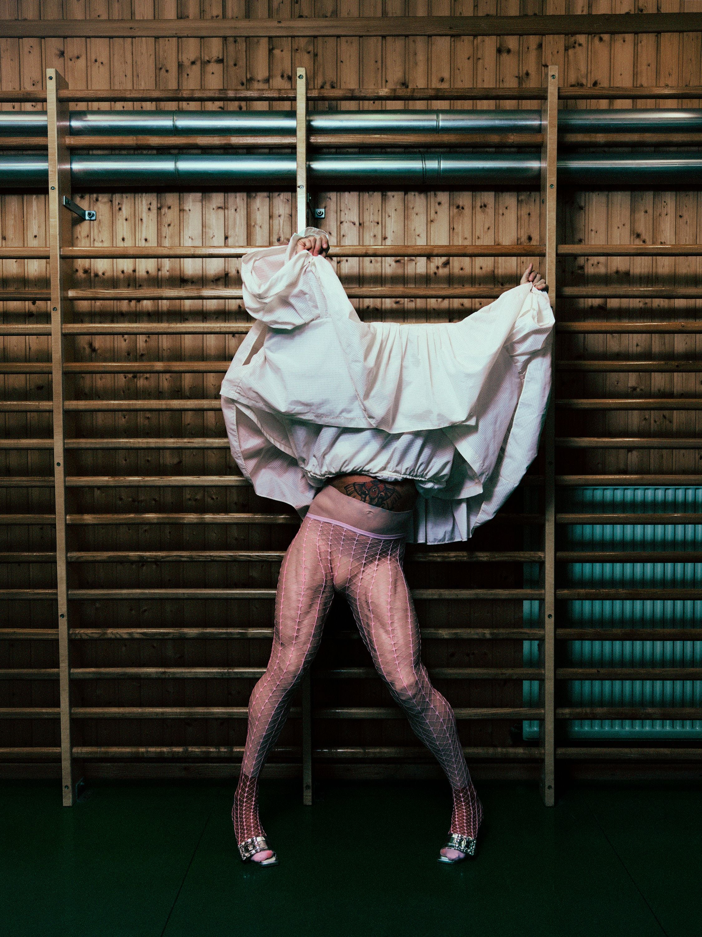Person in rosa Netzstrumpfhose und weißem, hochgezogenen Shirt an Sprossenwand in Turnhalle.