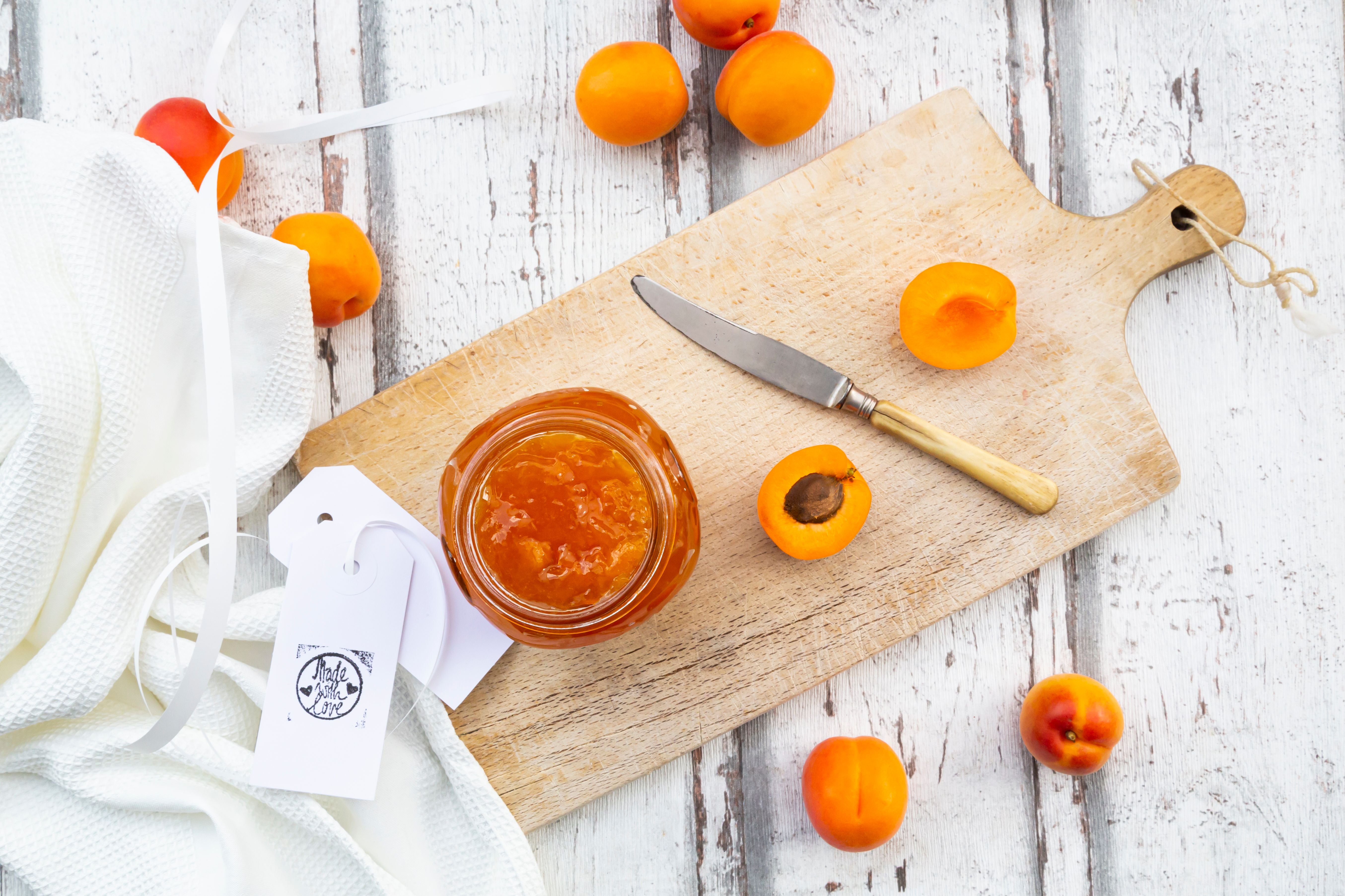 Ein Glas Marillenmarmelade mit frischen Marillen und Messer auf einem Holzbrett.