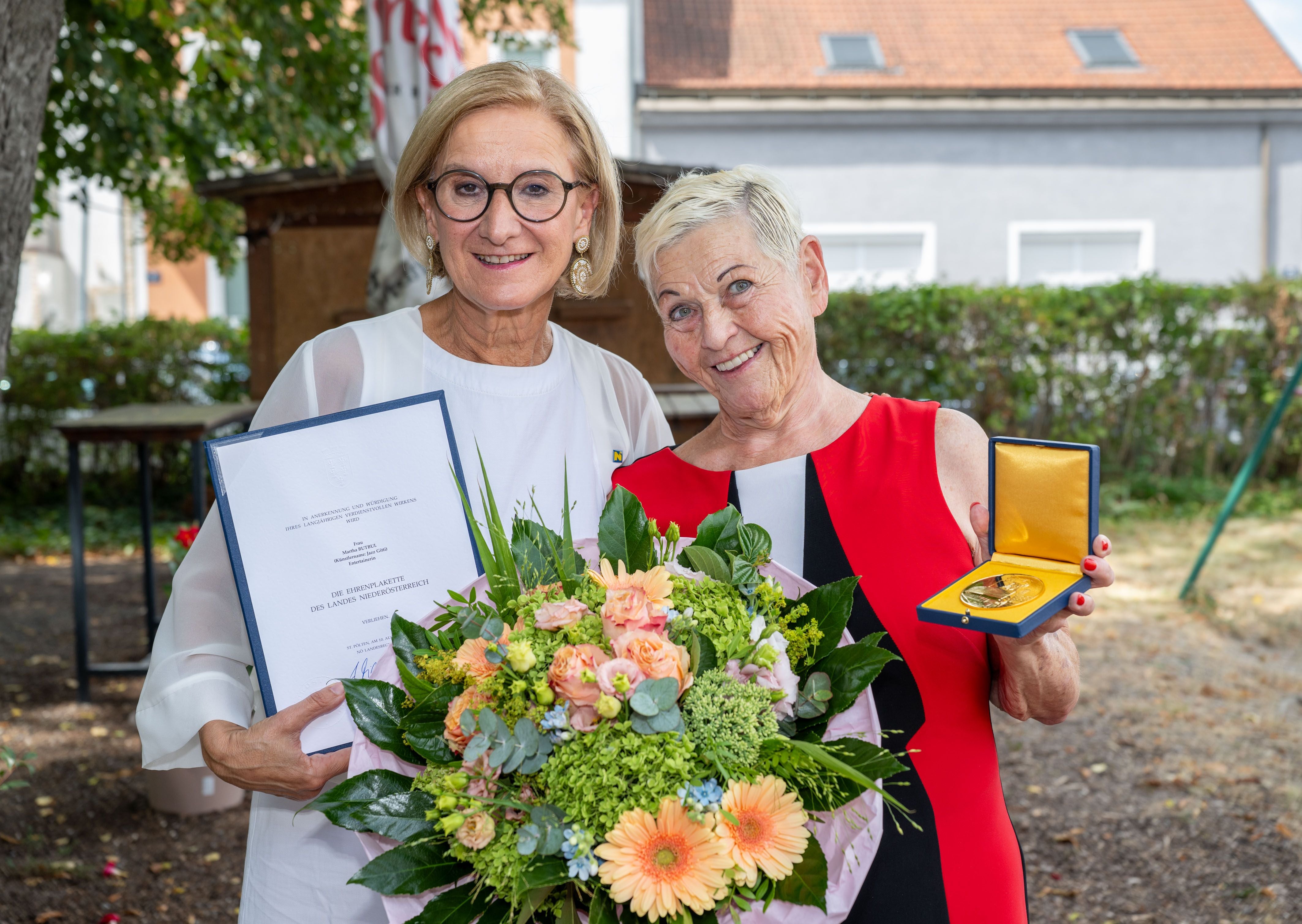 Zwei Frauen mit Urkunde, Blumenstrauß und goldener Ehrenplakette im Freien.
