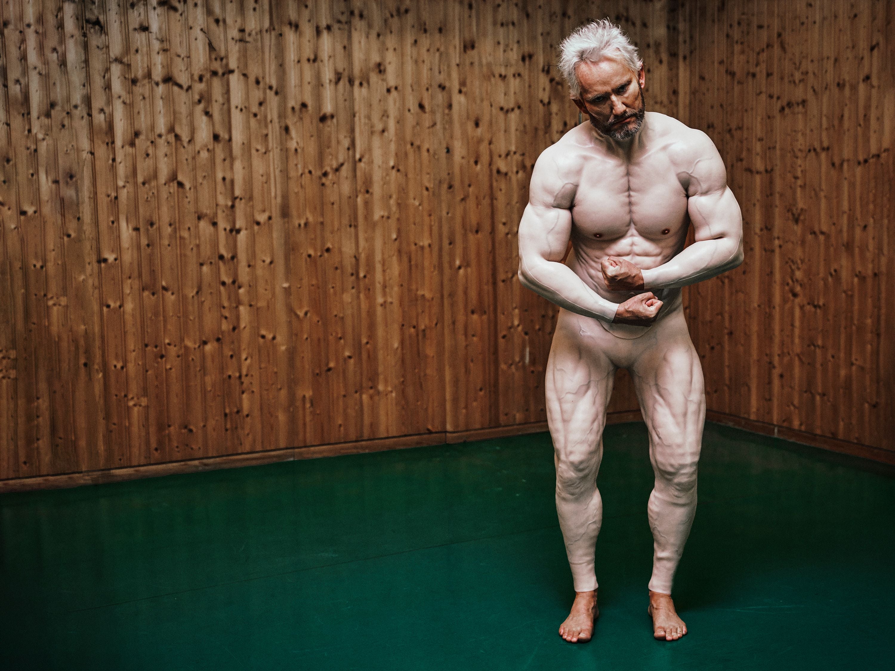 Muskelbepackter älterer Mann posiert im Bodybuilding-Stil vor einer Holzwand auf grünem Boden.