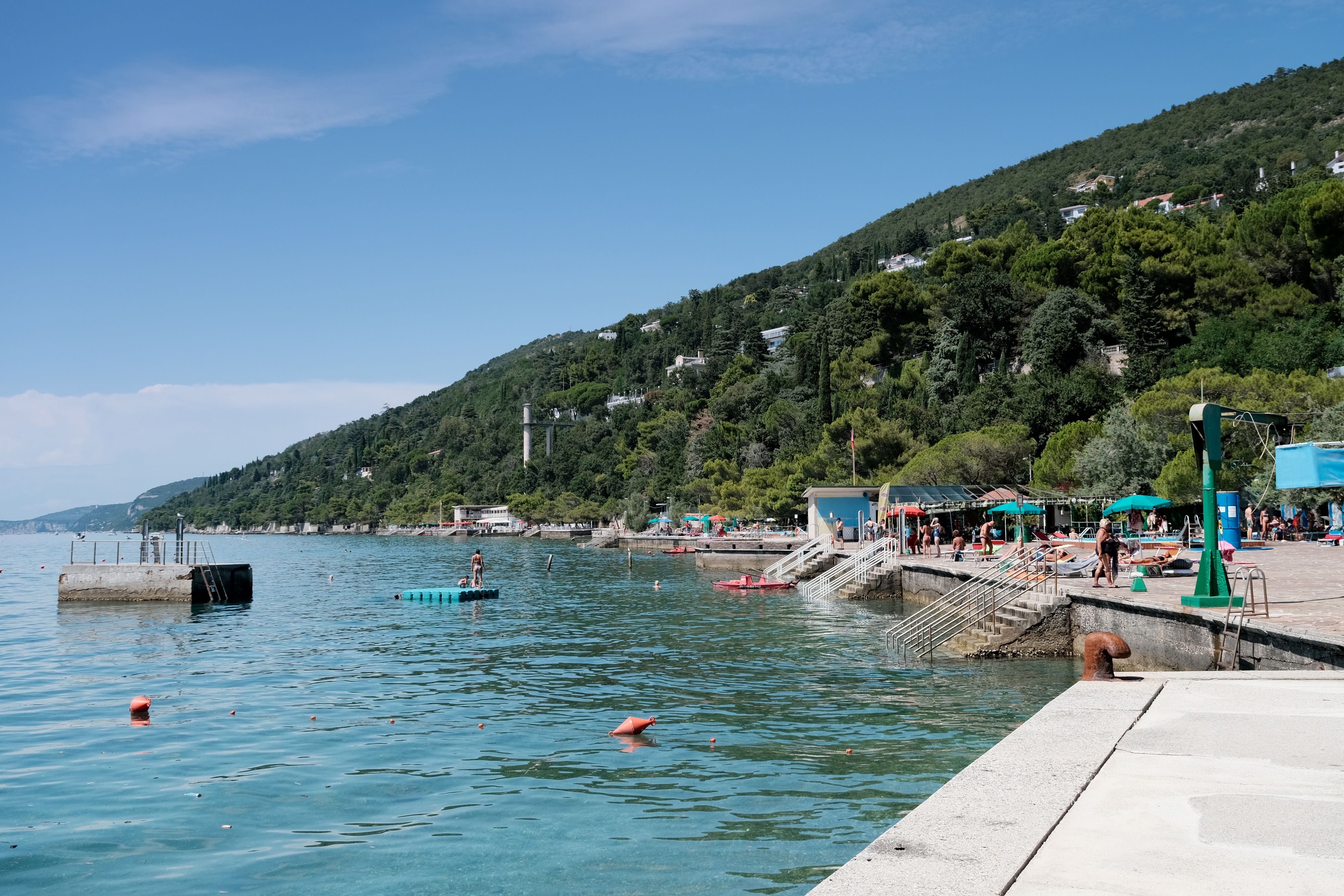 Menschen entspannen am Strand der Bucht Grignano bei Triest an der Adriaküste, umgeben von grünen Hügeln.