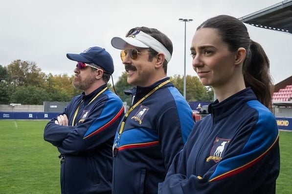 Drei Trainer im blauen Trainingsanzug stehen nebeneinander auf einem Fußballfeld.