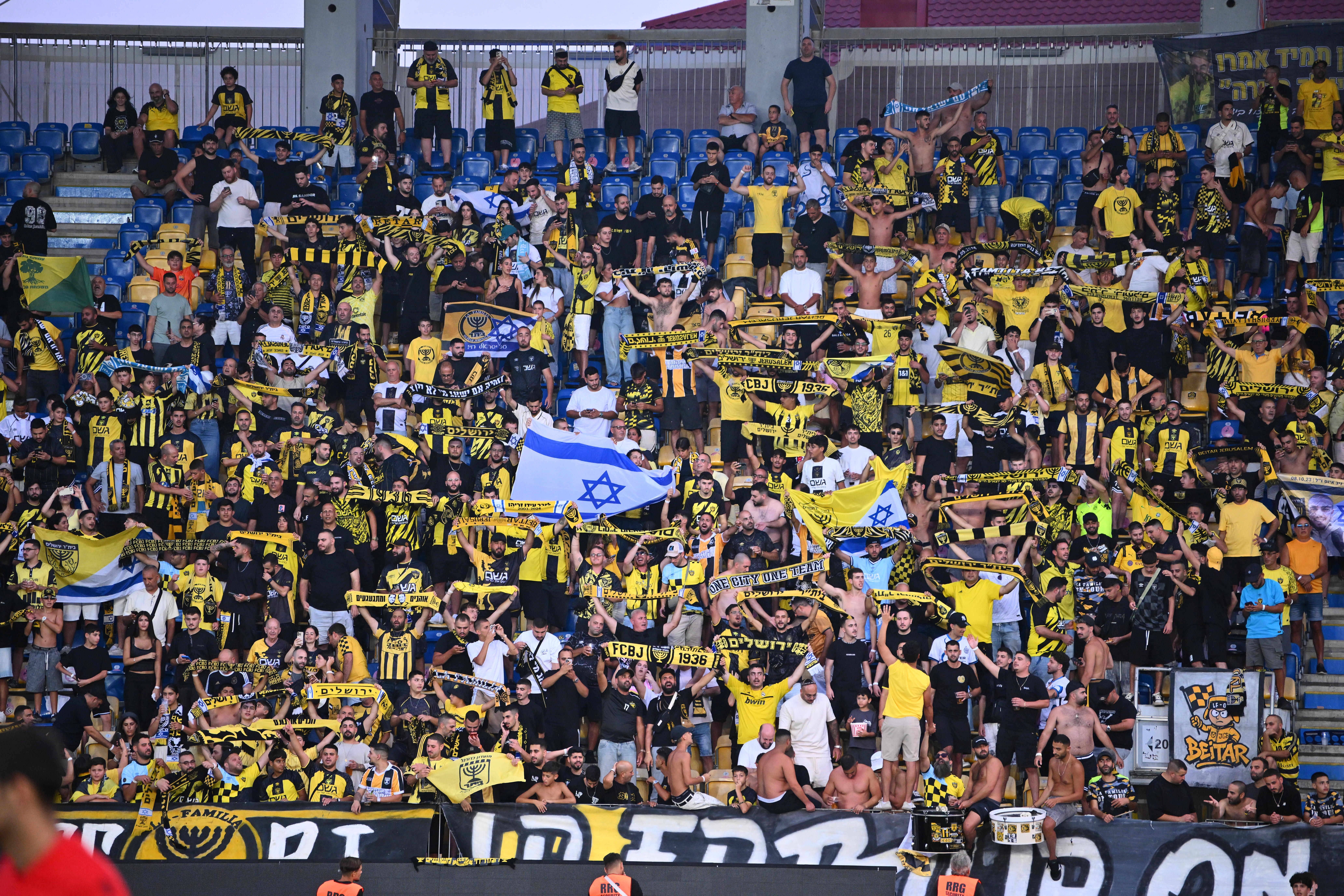 Fans von Beitar Jerusalem jubeln im Stadion, schwenken israelische Flaggen und gelb-schwarze Schals.