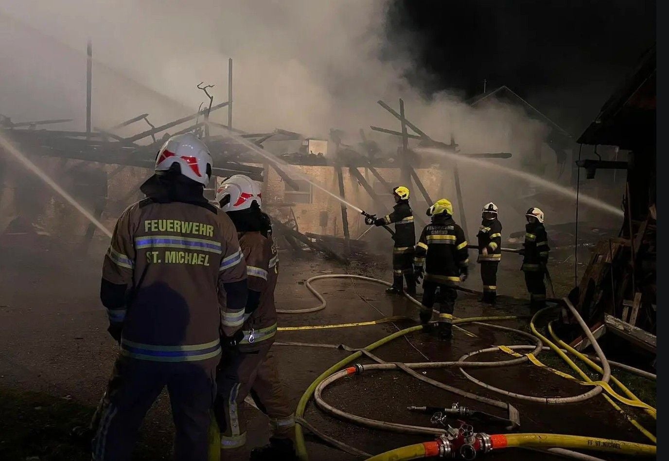 Feuerwehrleute bekämpfen nachts einen Brand mit Schläuchen, starker Rauch im Hintergrund.