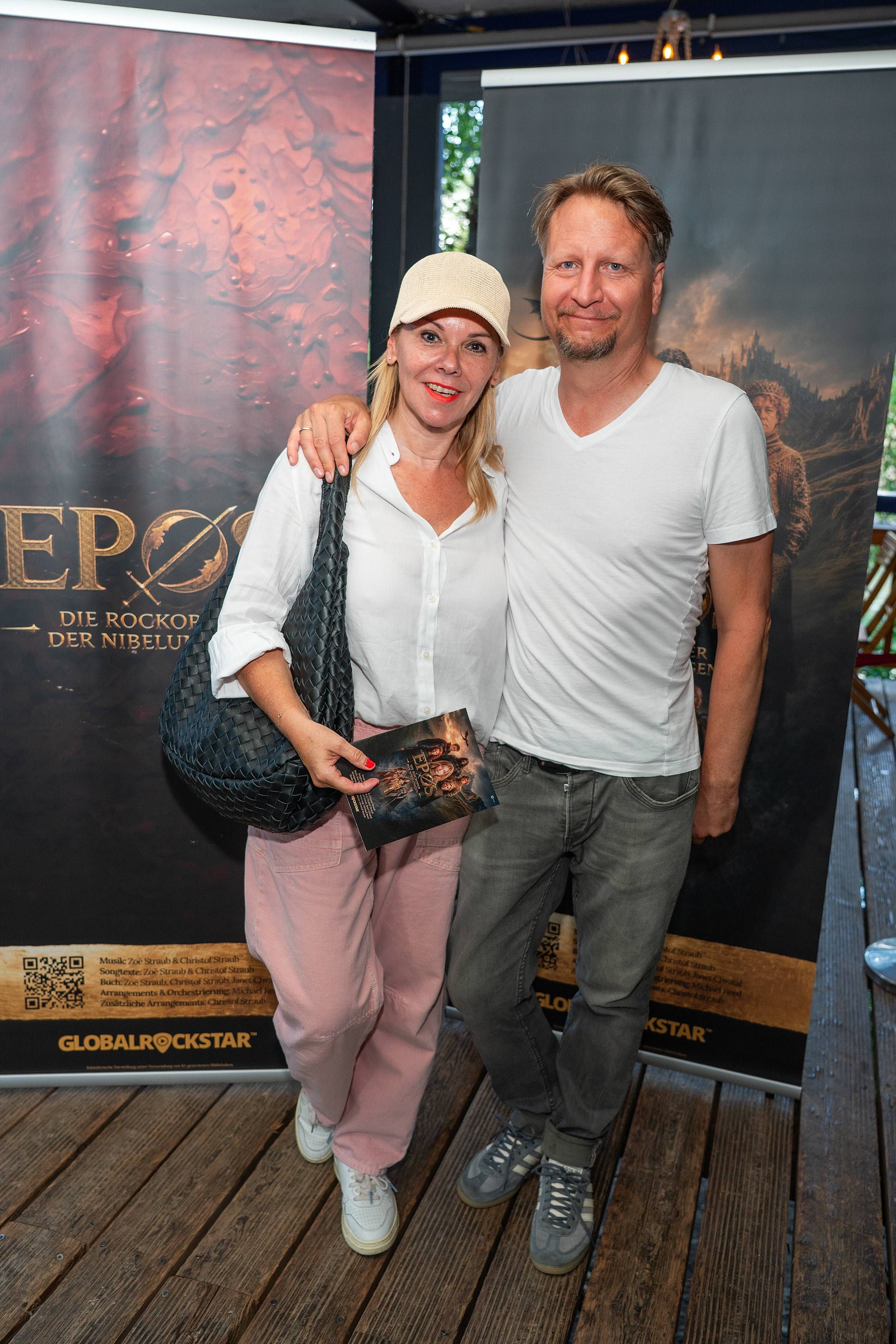 Ein Paar posiert lächelnd vor Plakaten zur Rockoper „EPOS“ bei einer Präsentation.