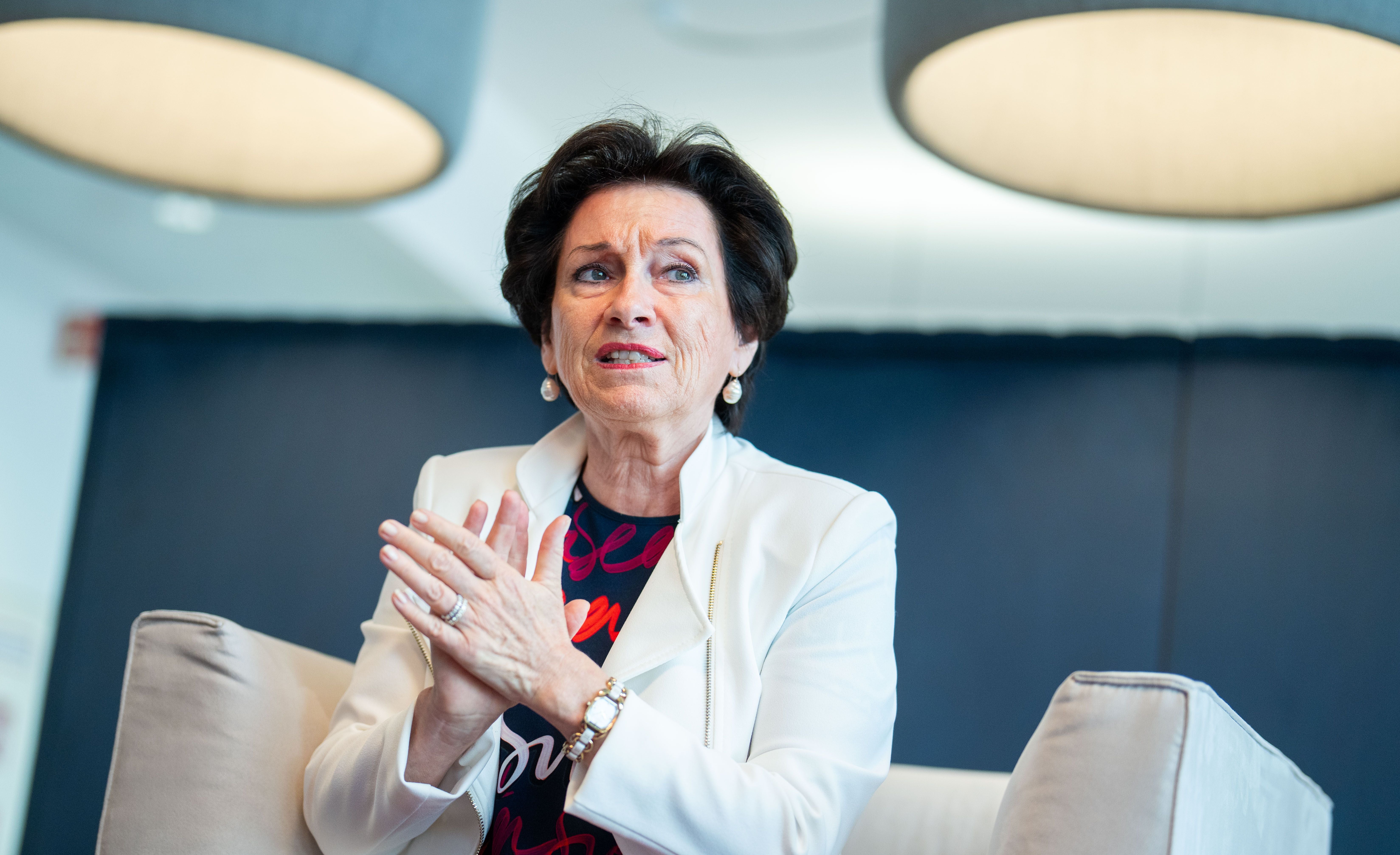 Frau im weißen Blazer spricht gestikulierend auf einem Sessel in modernem Büro.