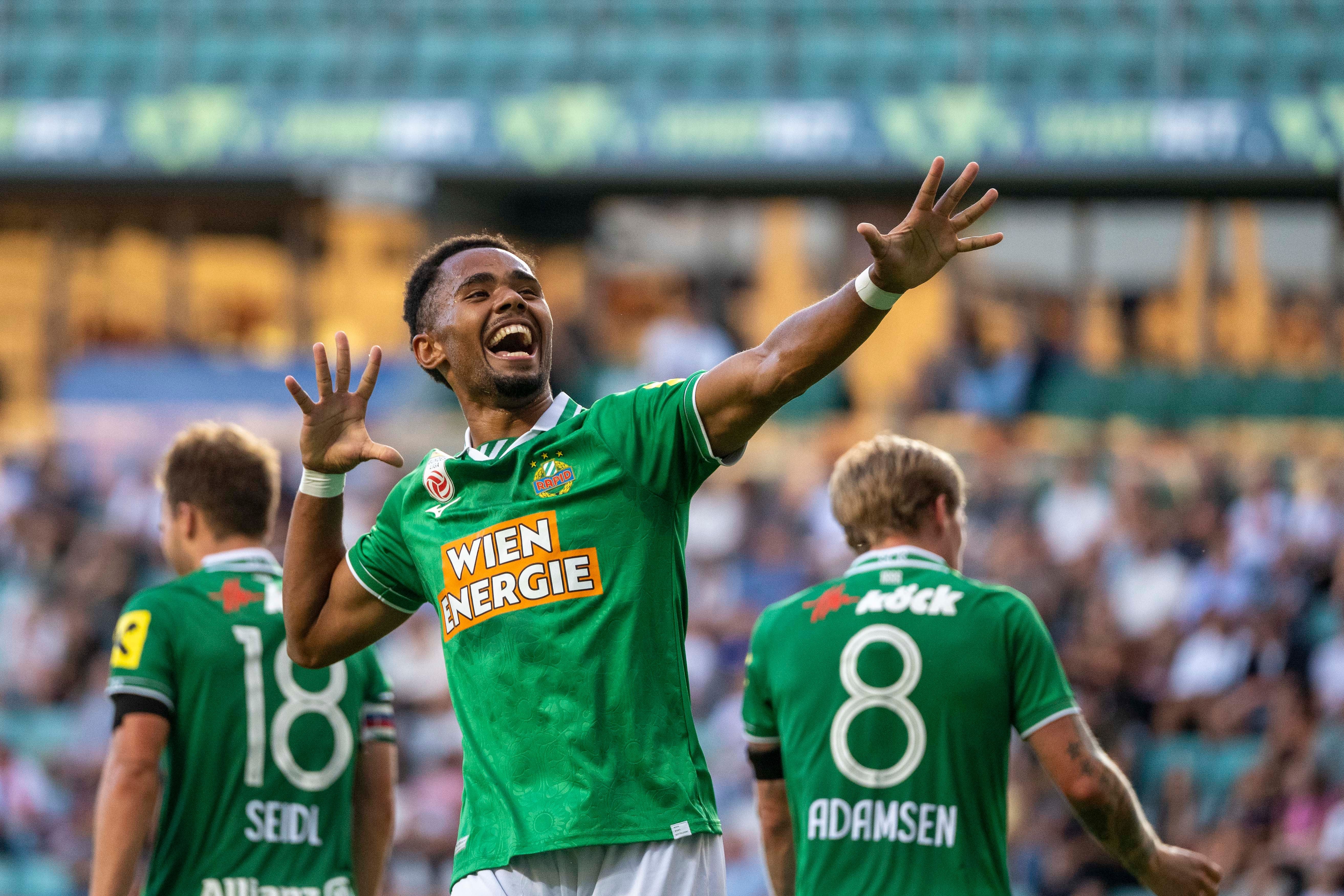 Ein Rapid Wien Spieler jubelt mit ausgestreckten Armen nach einem Tor, Teamkollegen im Hintergrund.