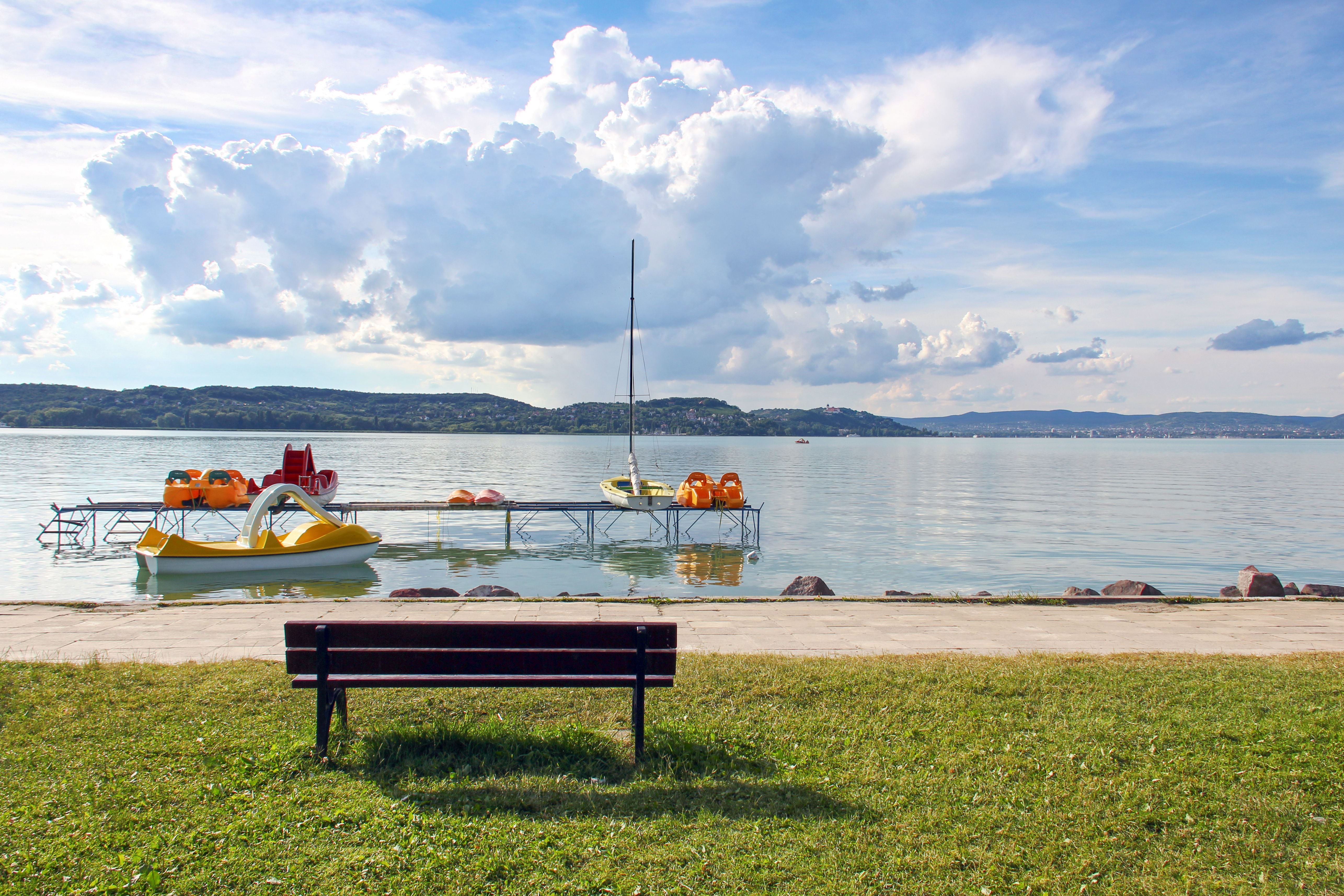 Eine Bank am Ufer mit Blick auf einen See, einen Steg und Tretboote bei sonnigem Wetter.