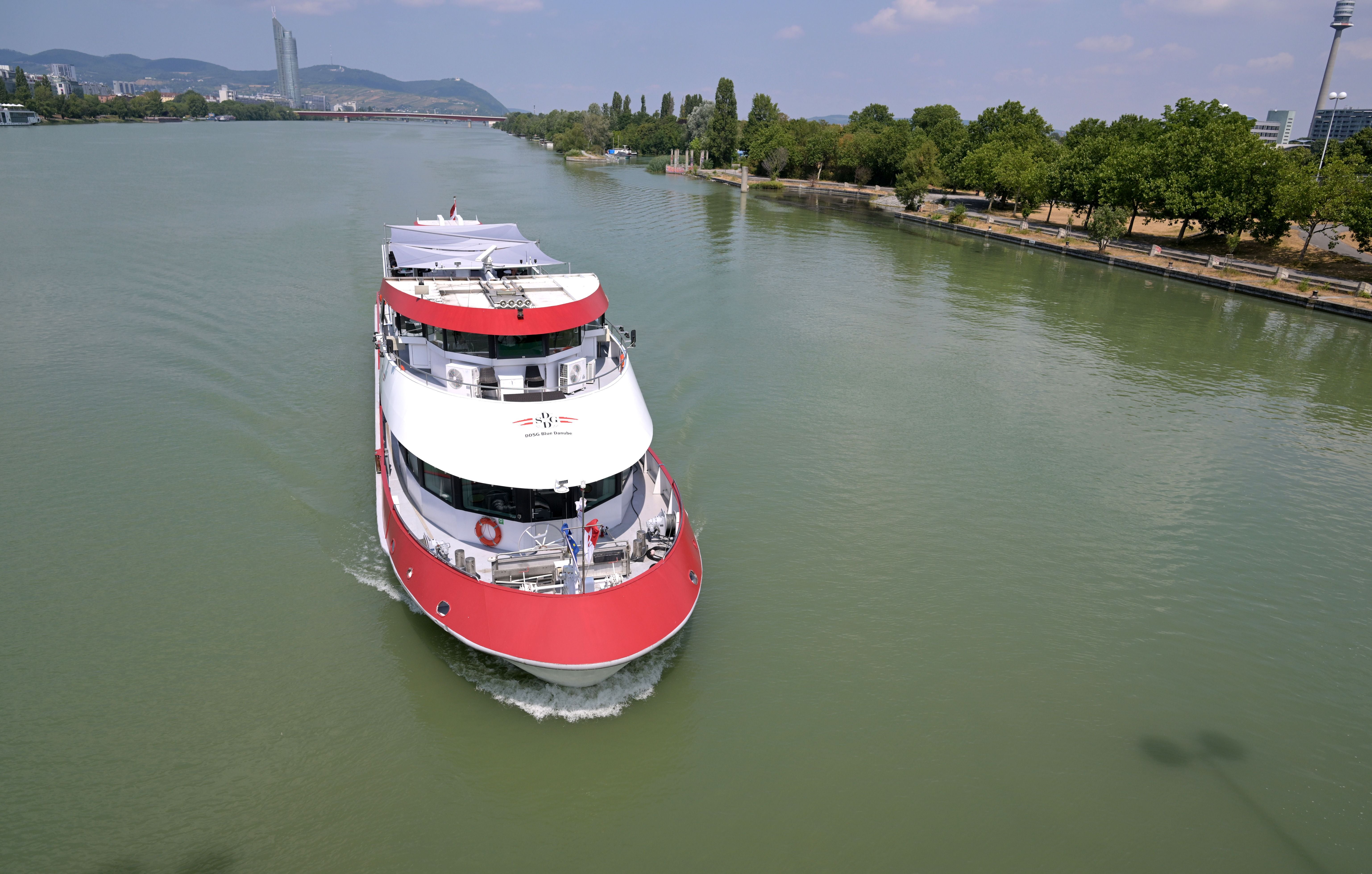 Ein Passagierschiff fährt auf der Donau in Wien entlang eines baumbestandenen Ufers.