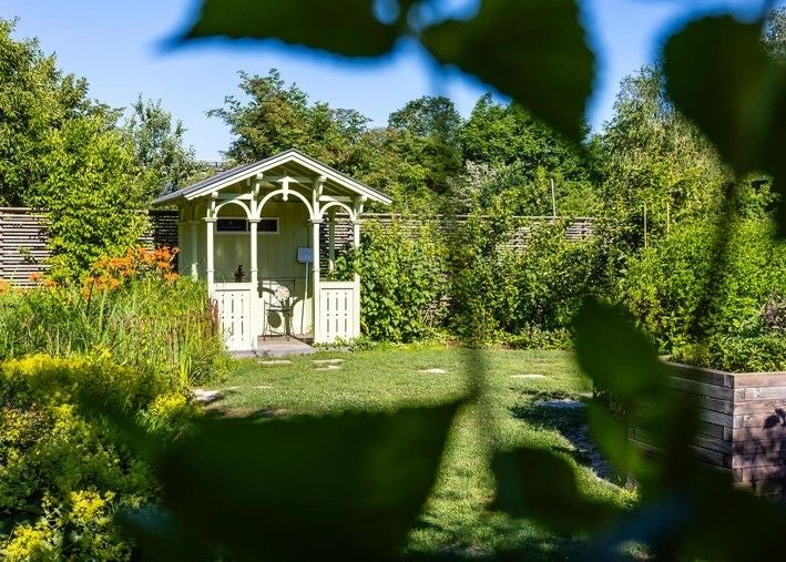 Ein hellgrünes Gartenhaus steht umgeben von üppigem Grün und Blumen im sonnigen Garten.