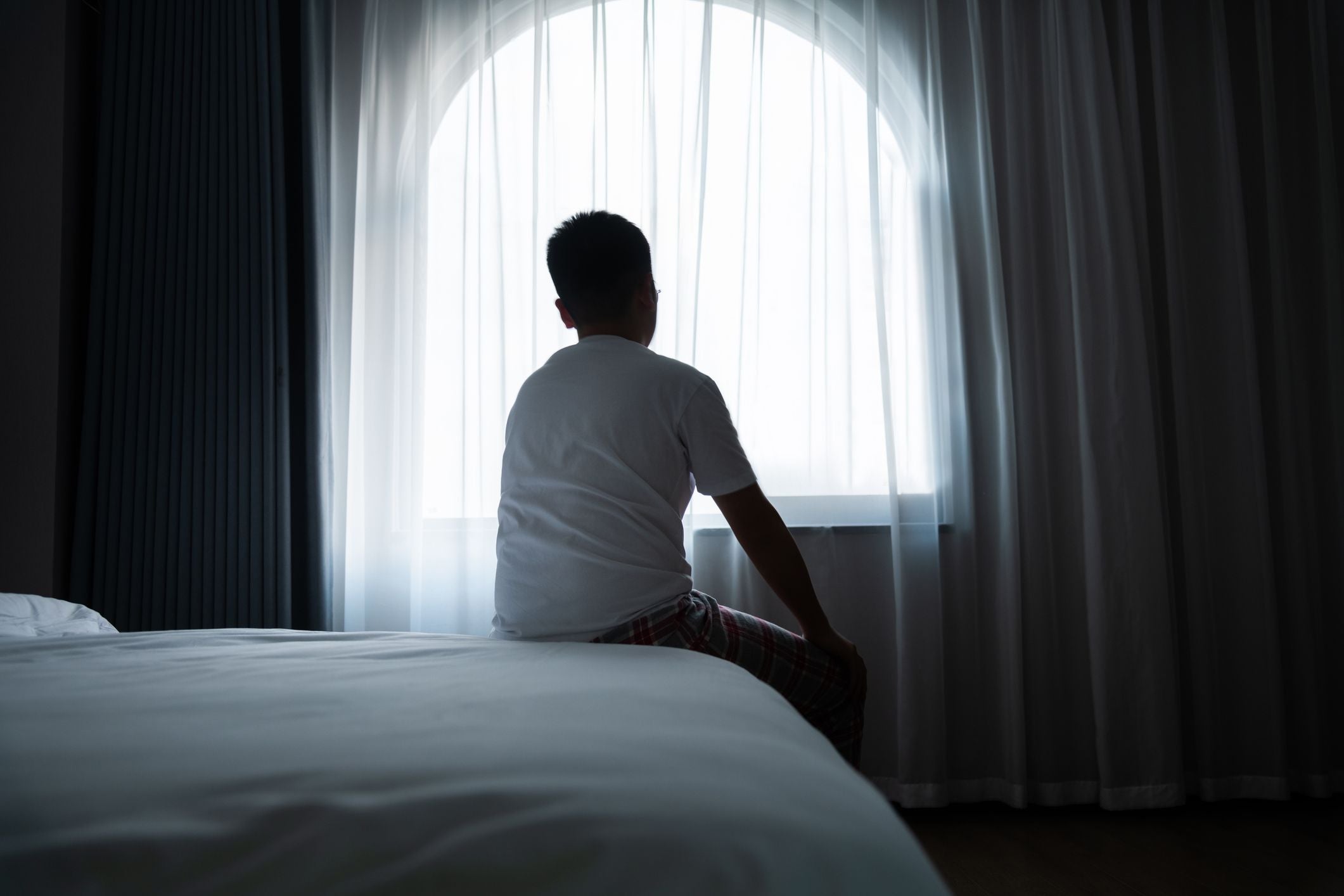 Ein Junge sitzt auf einem Bett und blickt durch ein Fenster mit transparentem Vorhang.