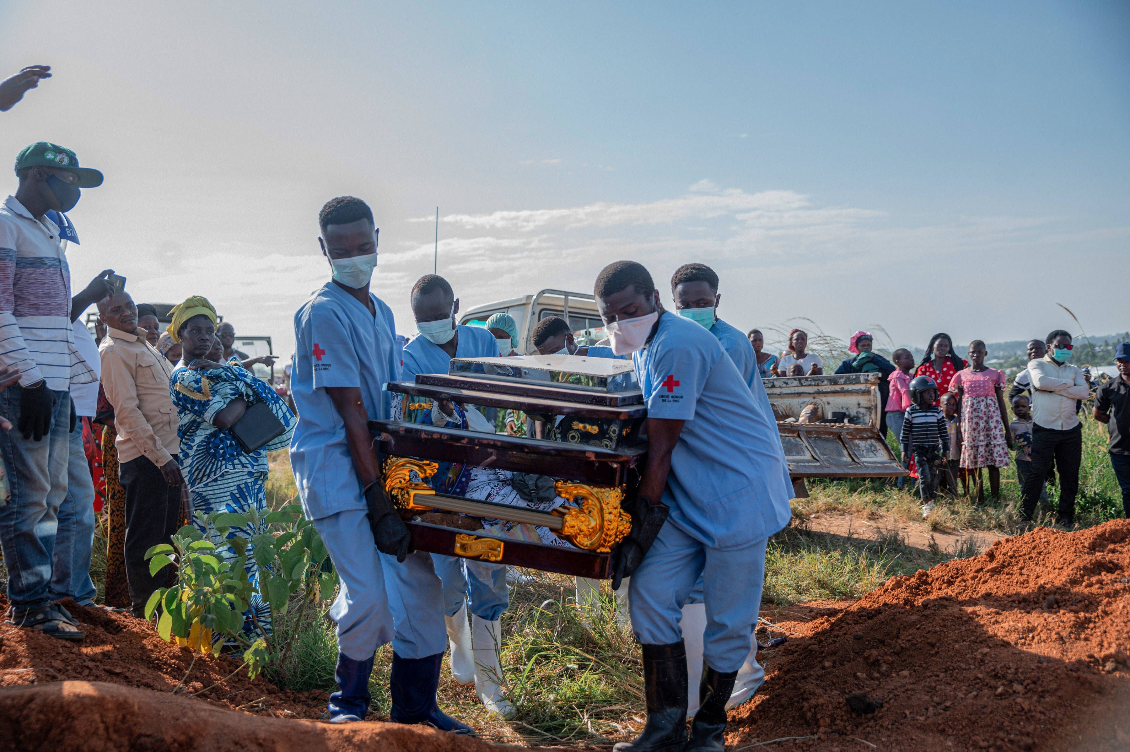 Gesundheitspersonal mit Schutzkleidung trägt einen Sarg, umgeben von trauernden Menschen auf einem Friedhof.