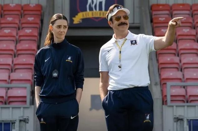 Zwei Trainer stehen auf einem Sportplatz, einer zeigt mit dem Arm nach vorne.