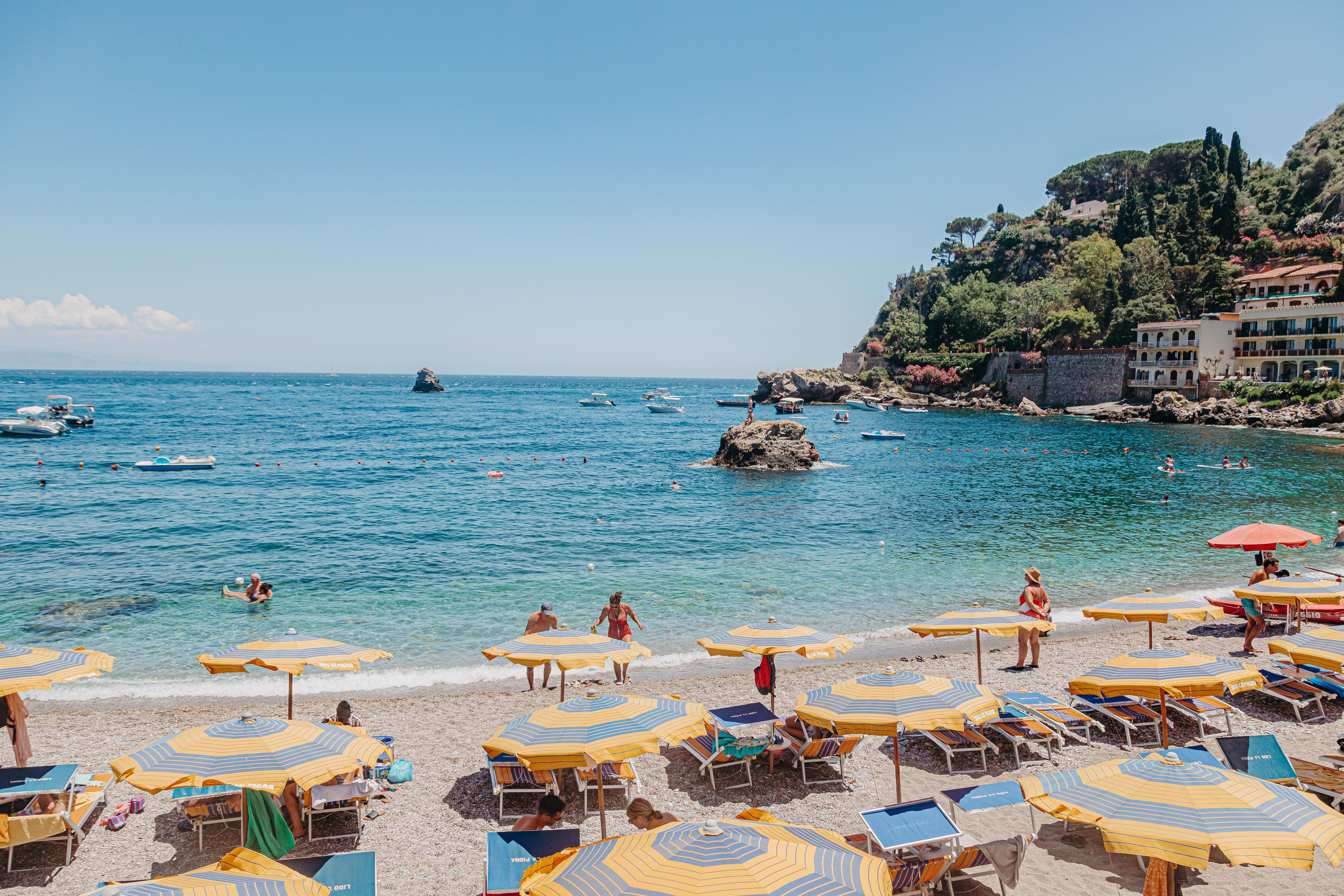 Strand mit gelb-blauen Sonnenschirmen, Liegestühlen und Badegästen am klaren Meer bei Taormina.
