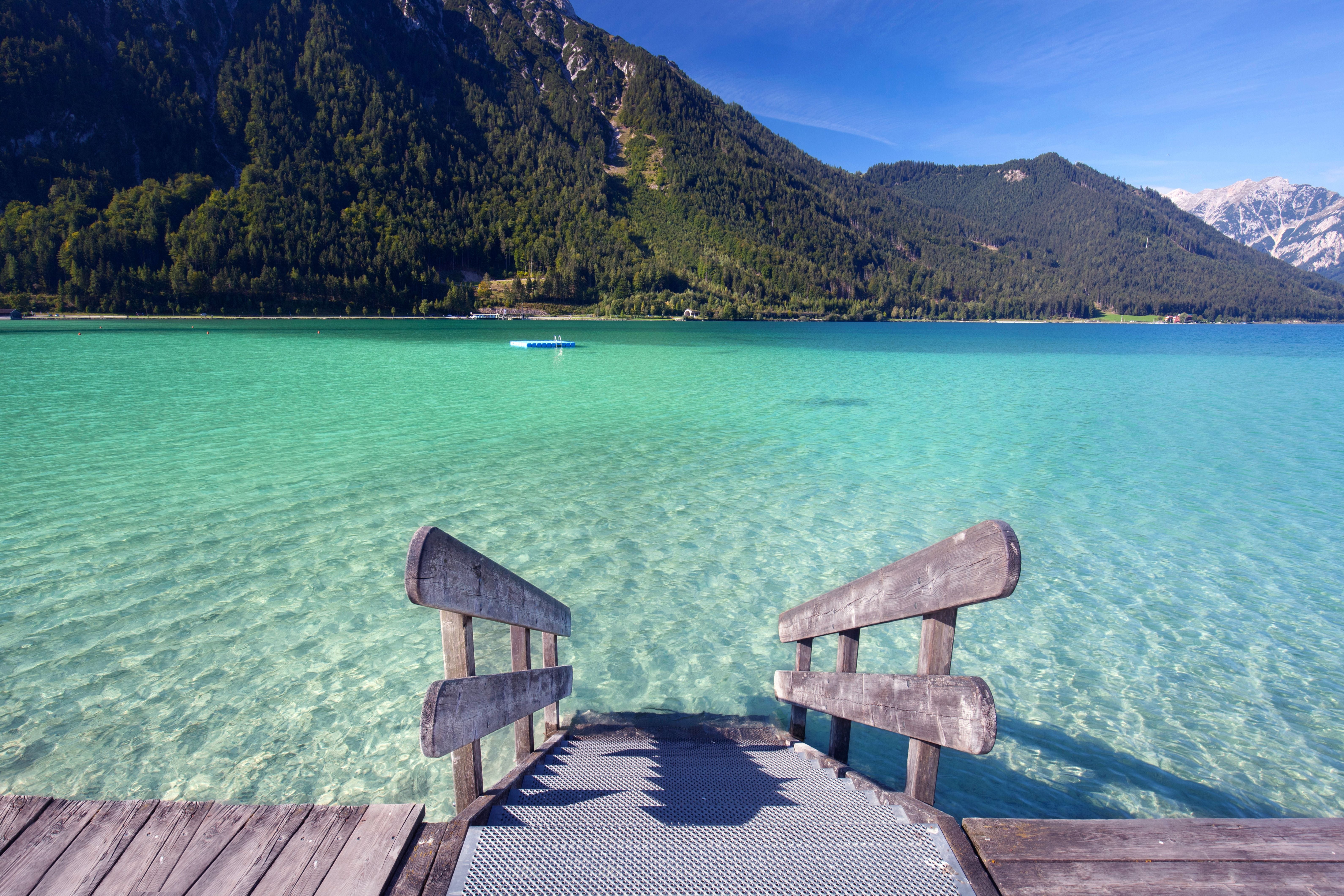 Steg mit Treppe führt in türkisfarbenen, klaren See vor bewaldeten Bergen und blauem Himmel.