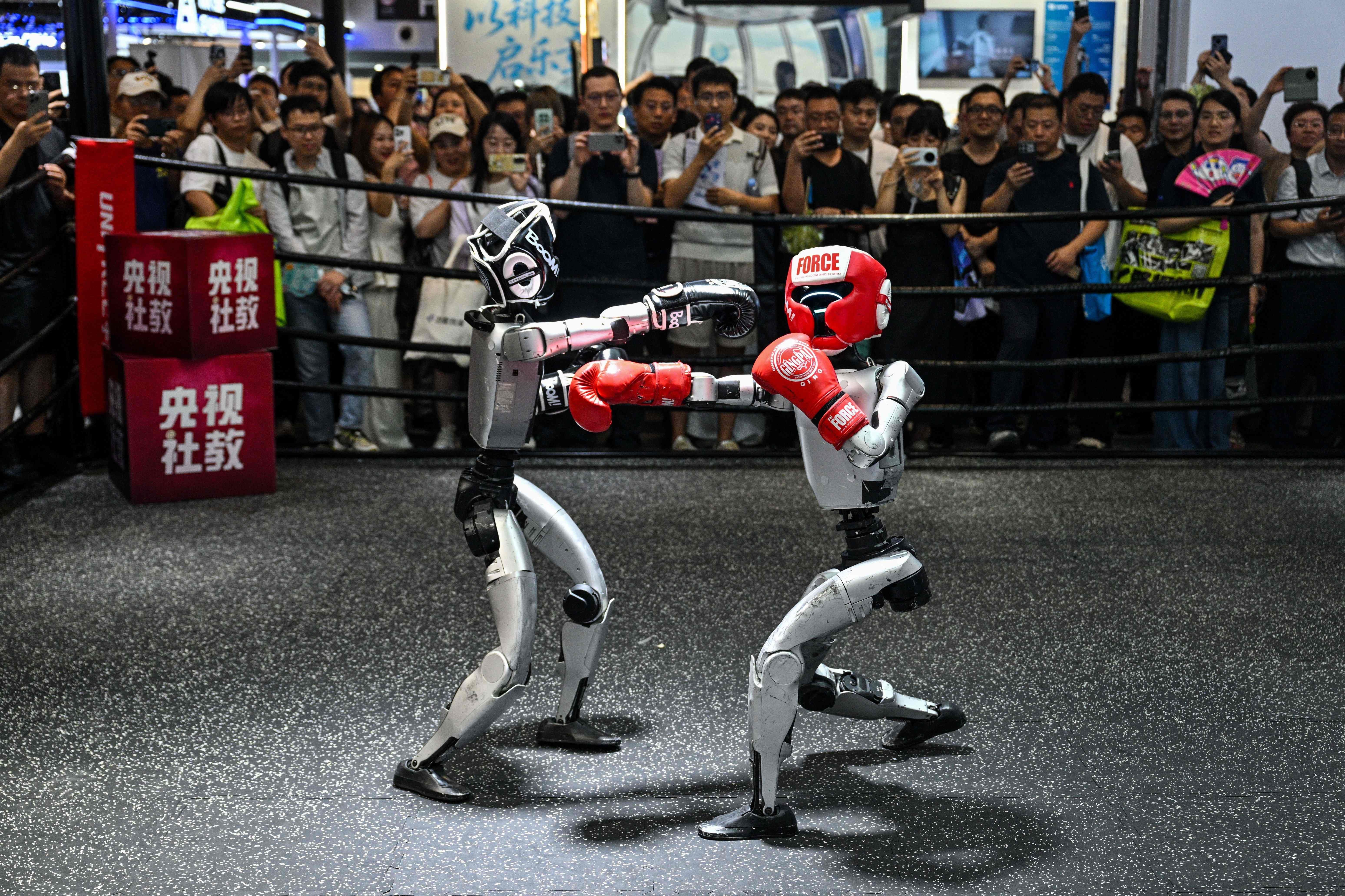 Zwei humanoide Roboter boxen im Ring vor einer zuschauenden Menschenmenge auf einer Messe.