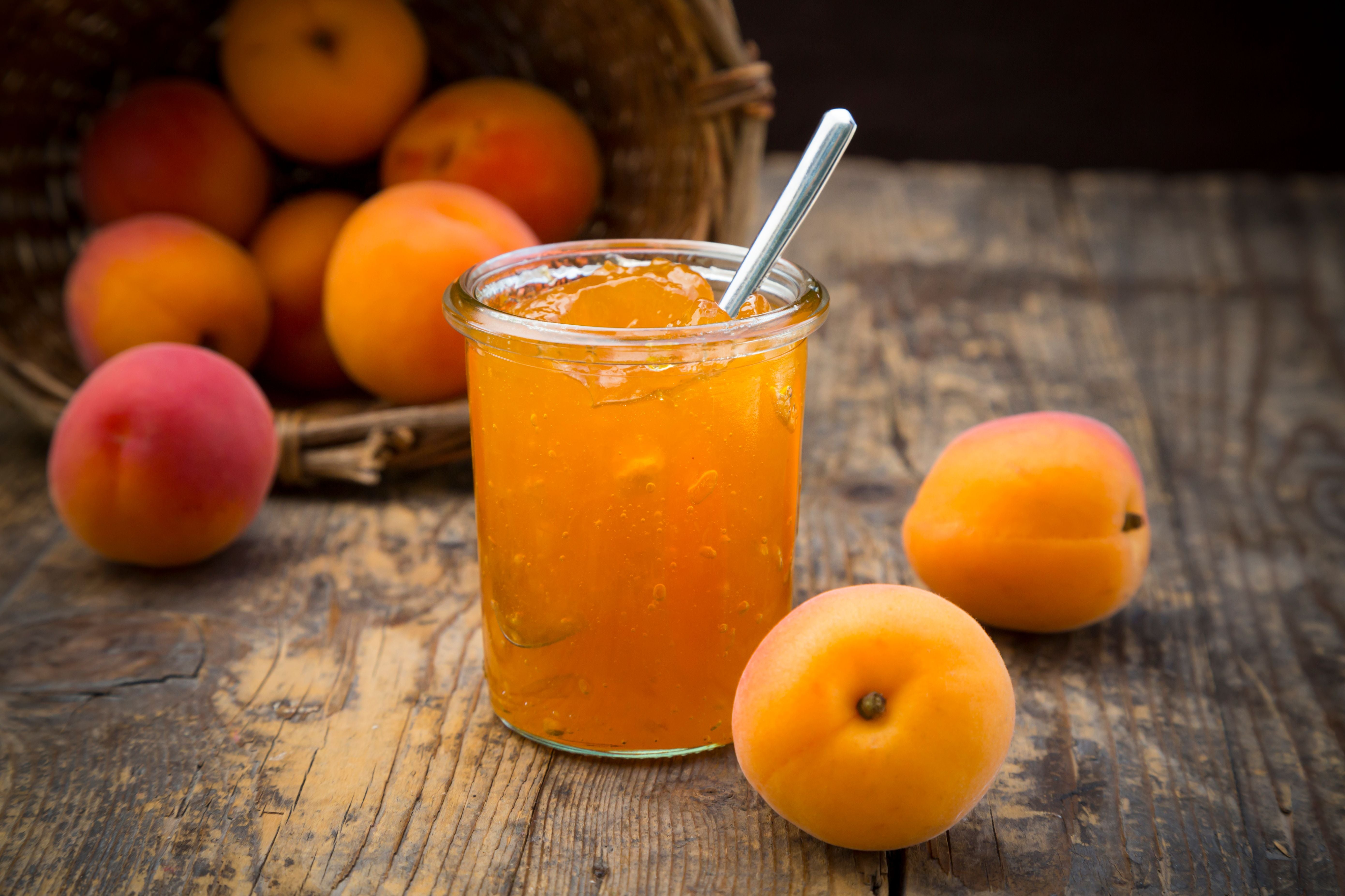 Ein Glas Marillenmarmelade mit Löffel, umgeben von frischen Marillen auf Holztisch.