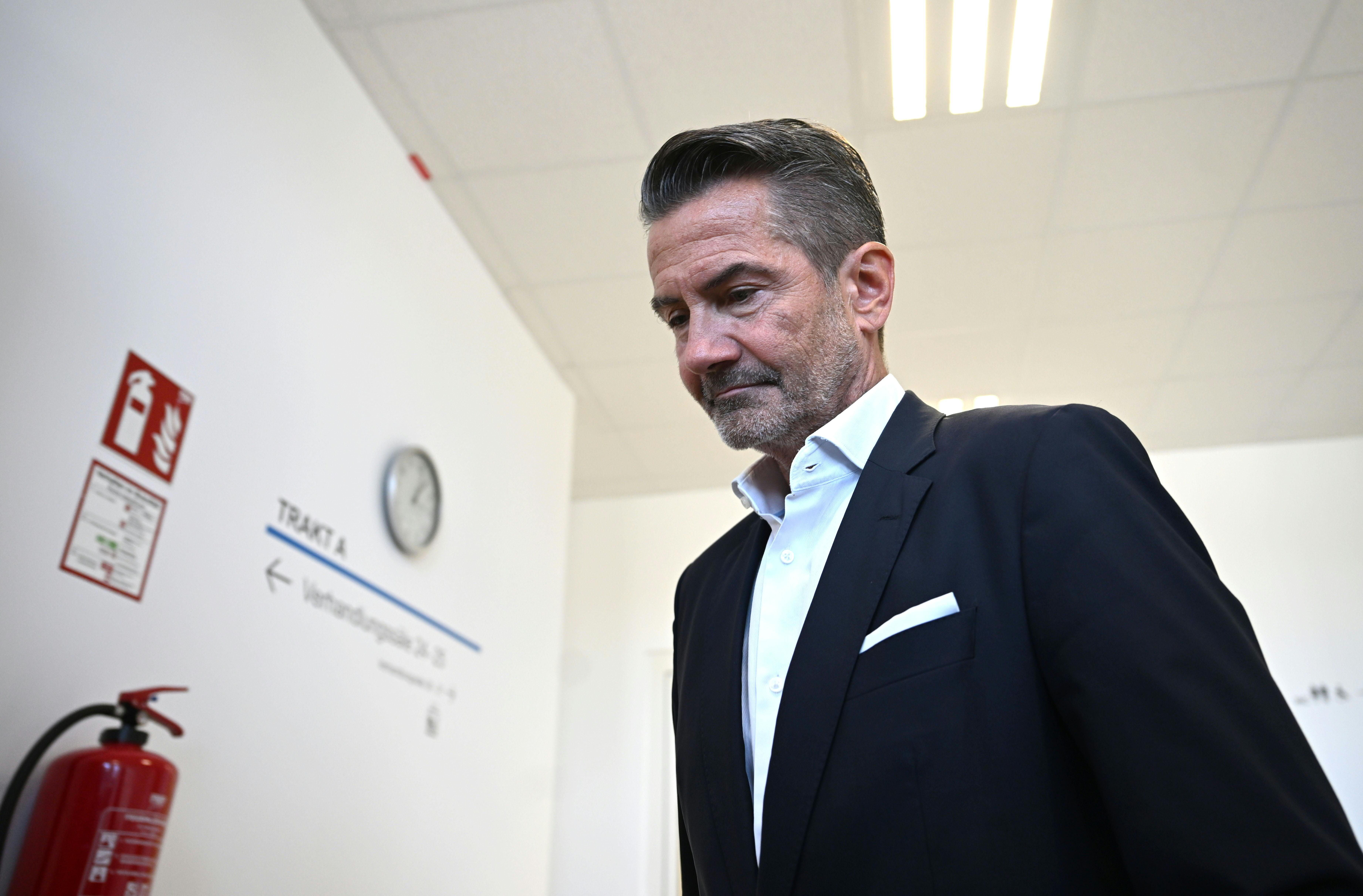 Ein Mann im Anzug steht nachdenklich in einem hellen Flur vor einer weißen Wand mit Uhr.
