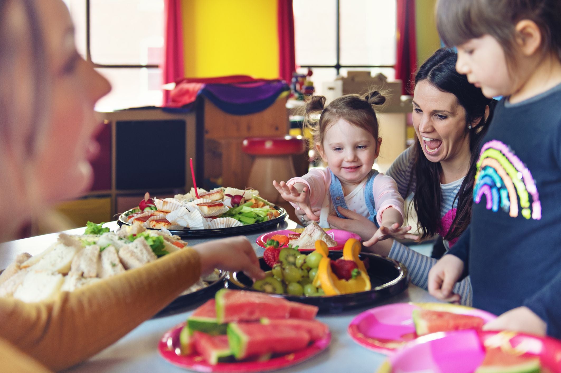Kinder essen im Kindergarten gemeinsam Obst und Sandwiches an einem bunt gedeckten Tisch.