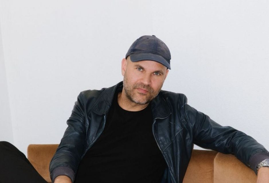 Mann mit schwarzer Lederjacke und dunkler Mütze sitzt auf einem braunen Sofa vor weißer Wand.