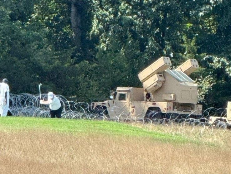 US-Präsident Trump spielt Golf neben einem Militärfahrzeug mit Flugabwehrraketen und Stacheldraht.