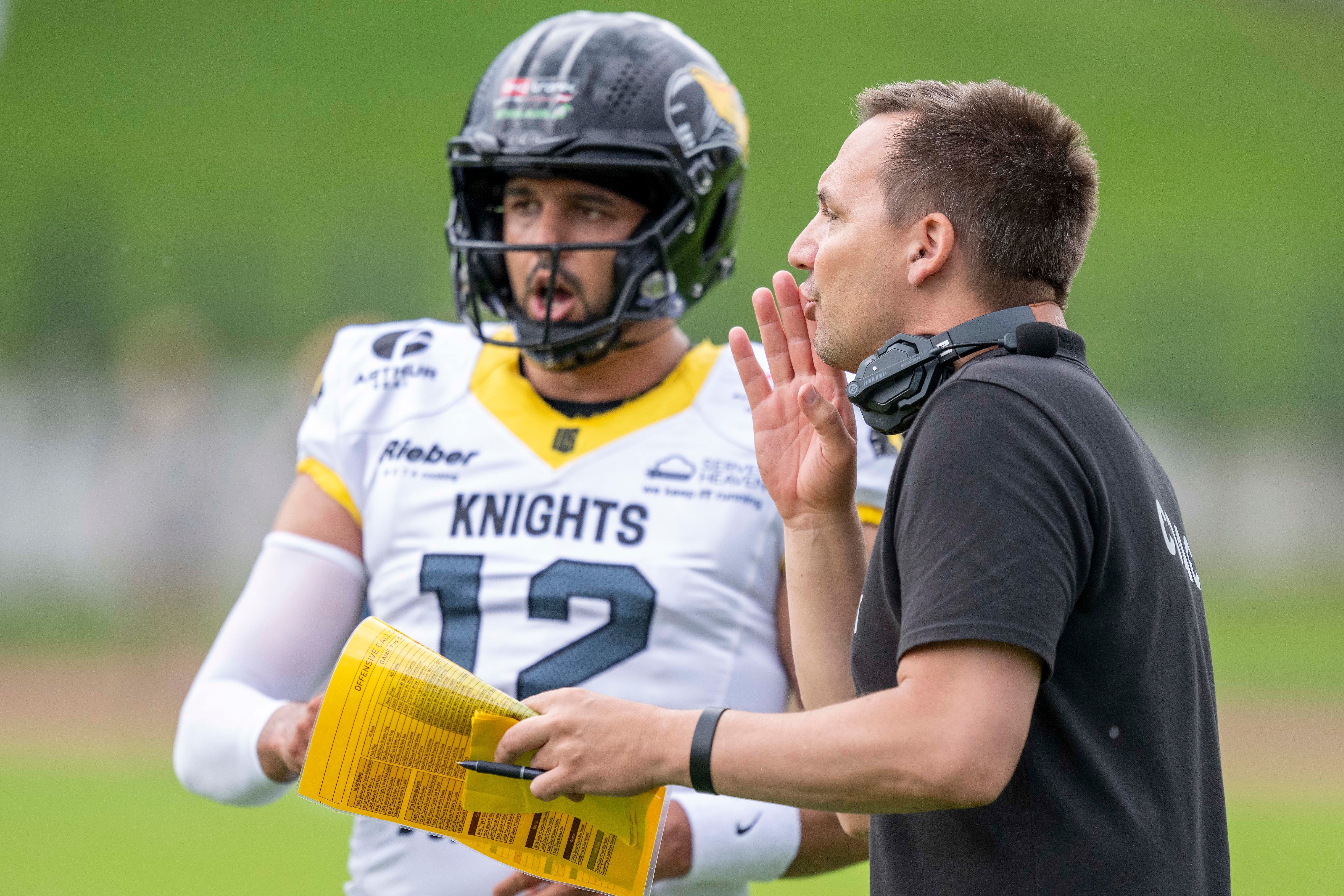 Footballtrainer spricht mit Spieler im Helm während eines Spiels, beide im Knights-Team-Outfit.