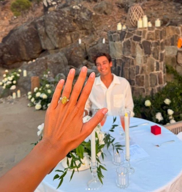 Eine Hand mit Verlobungsring im Vordergrund, dahinter ein lächelnder Mann an einem Tisch.