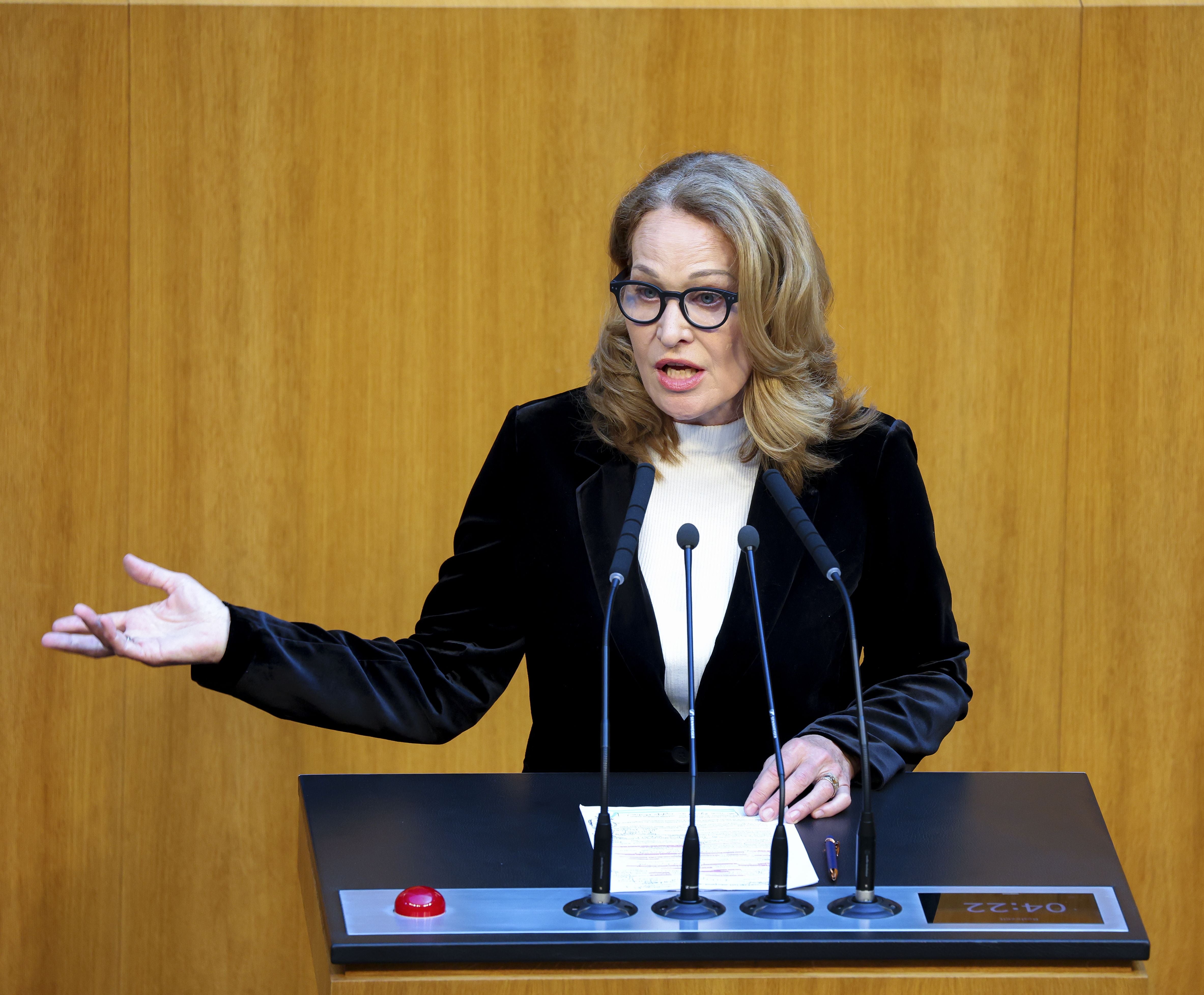 Frau spricht an einem Rednerpult im österreichischen Nationalrat, Holzvertäfelung im Hintergrund.