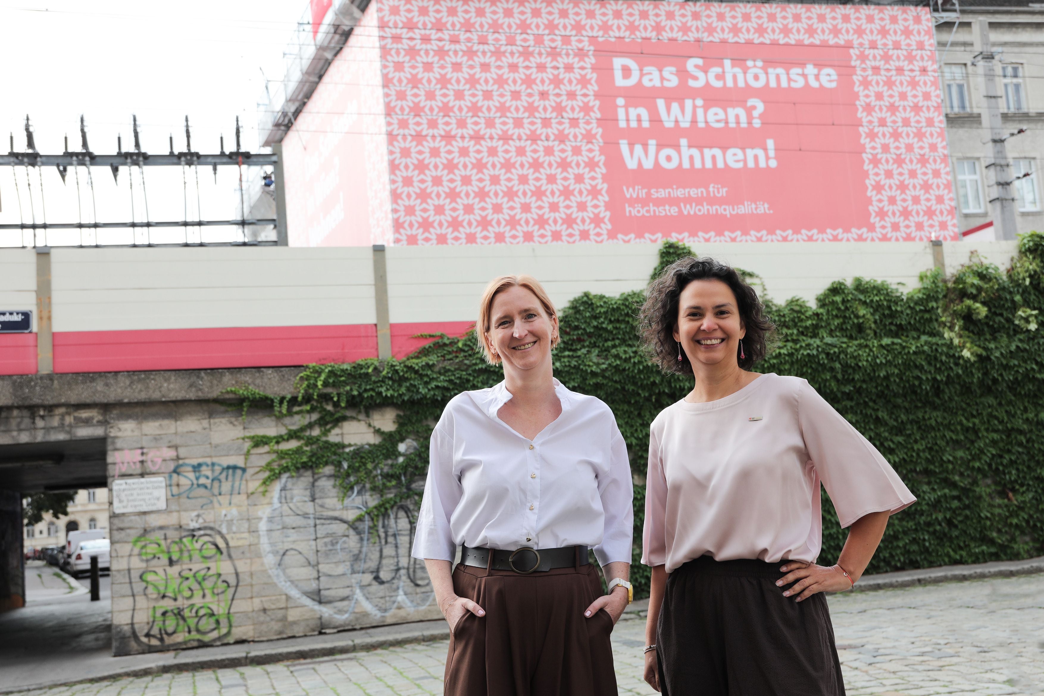 Zwei Frauen stehen lächelnd vor einer Baustelle mit großem roten Wohnplakat in Wien.