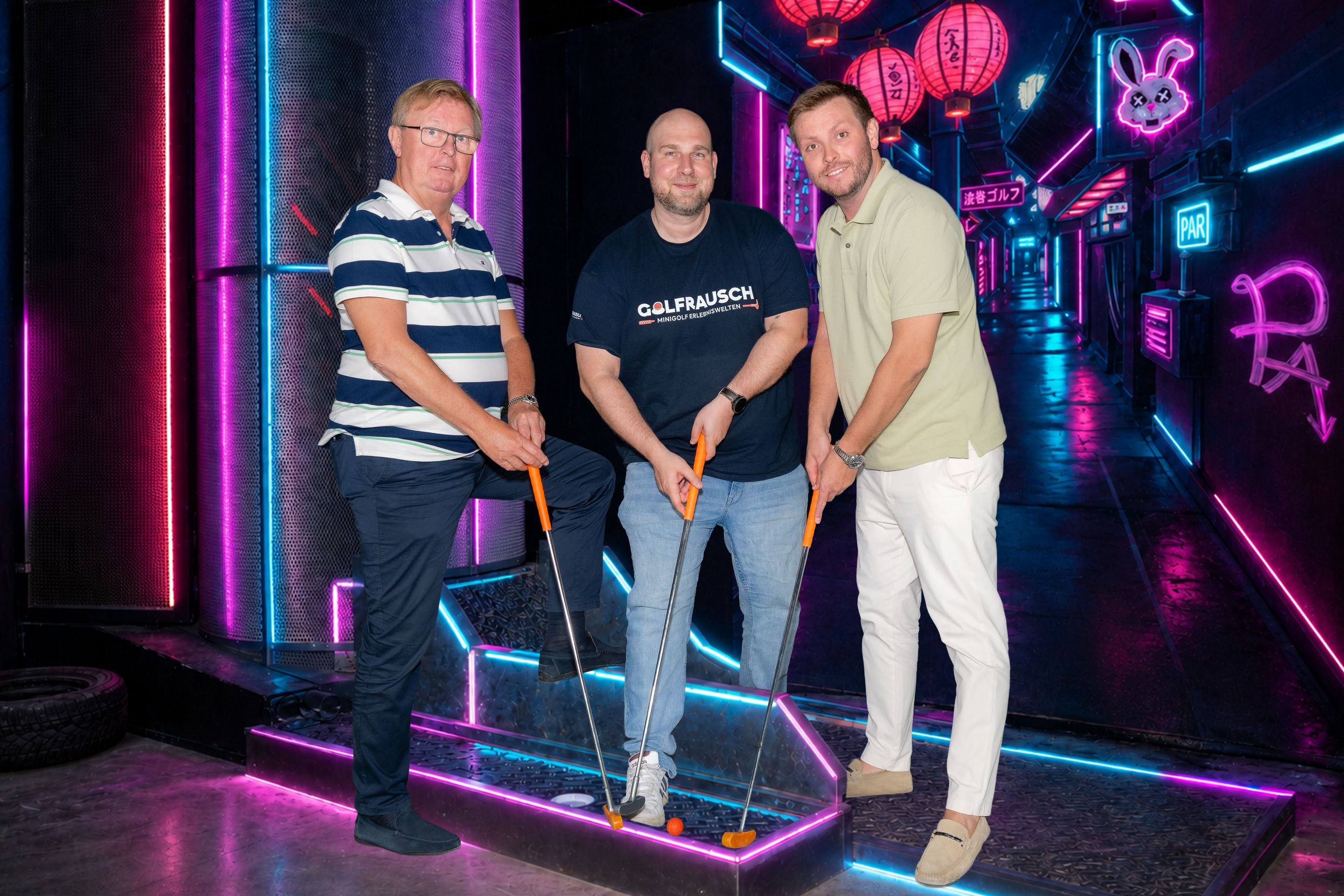 Drei Männer spielen Minigolf in einer neonbeleuchteten Indoorhalle mit futuristischer Dekoration.