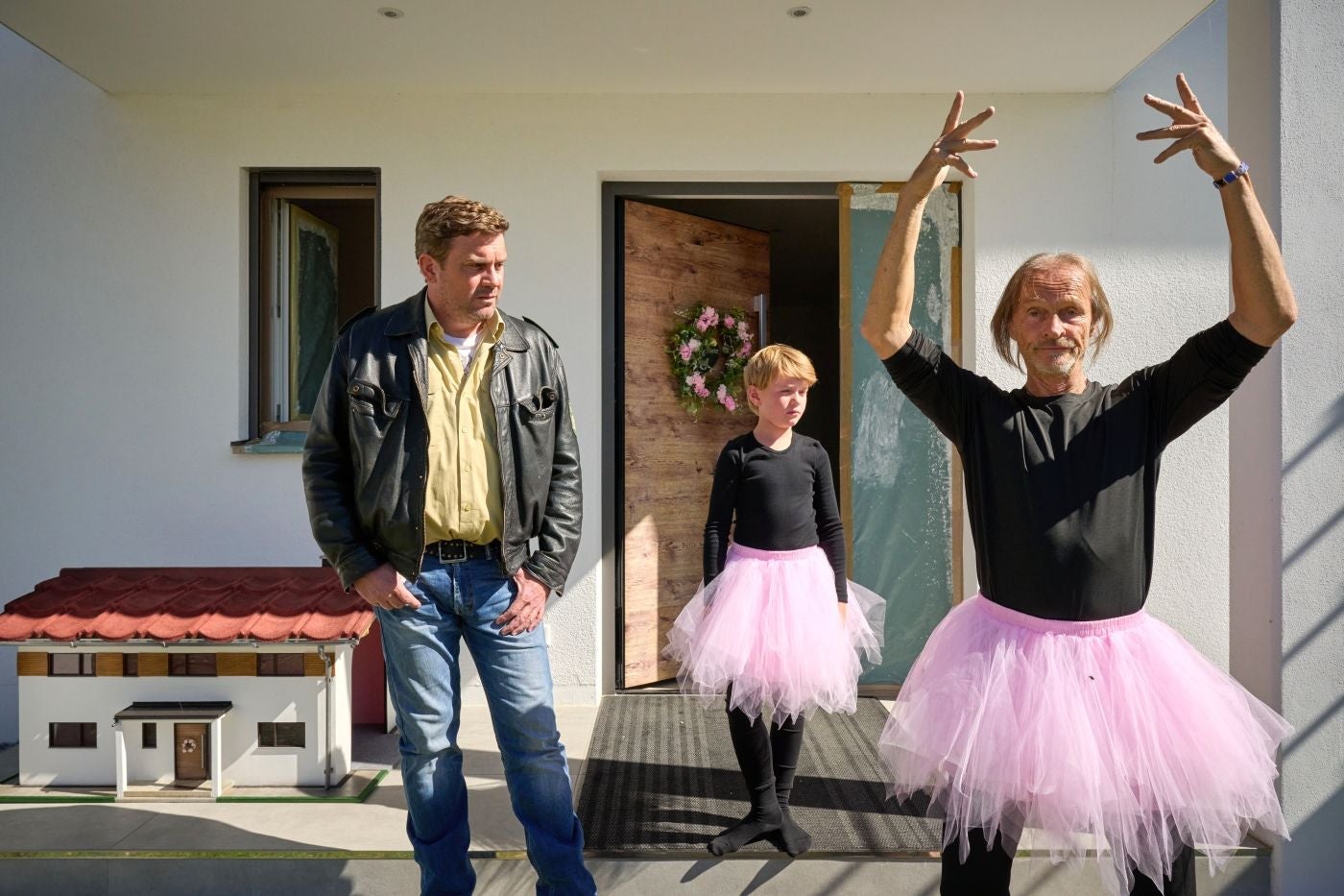 Ein Mann und ein Kind tragen rosa Tutu, ein weiterer Mann steht daneben auf einer Terrasse.