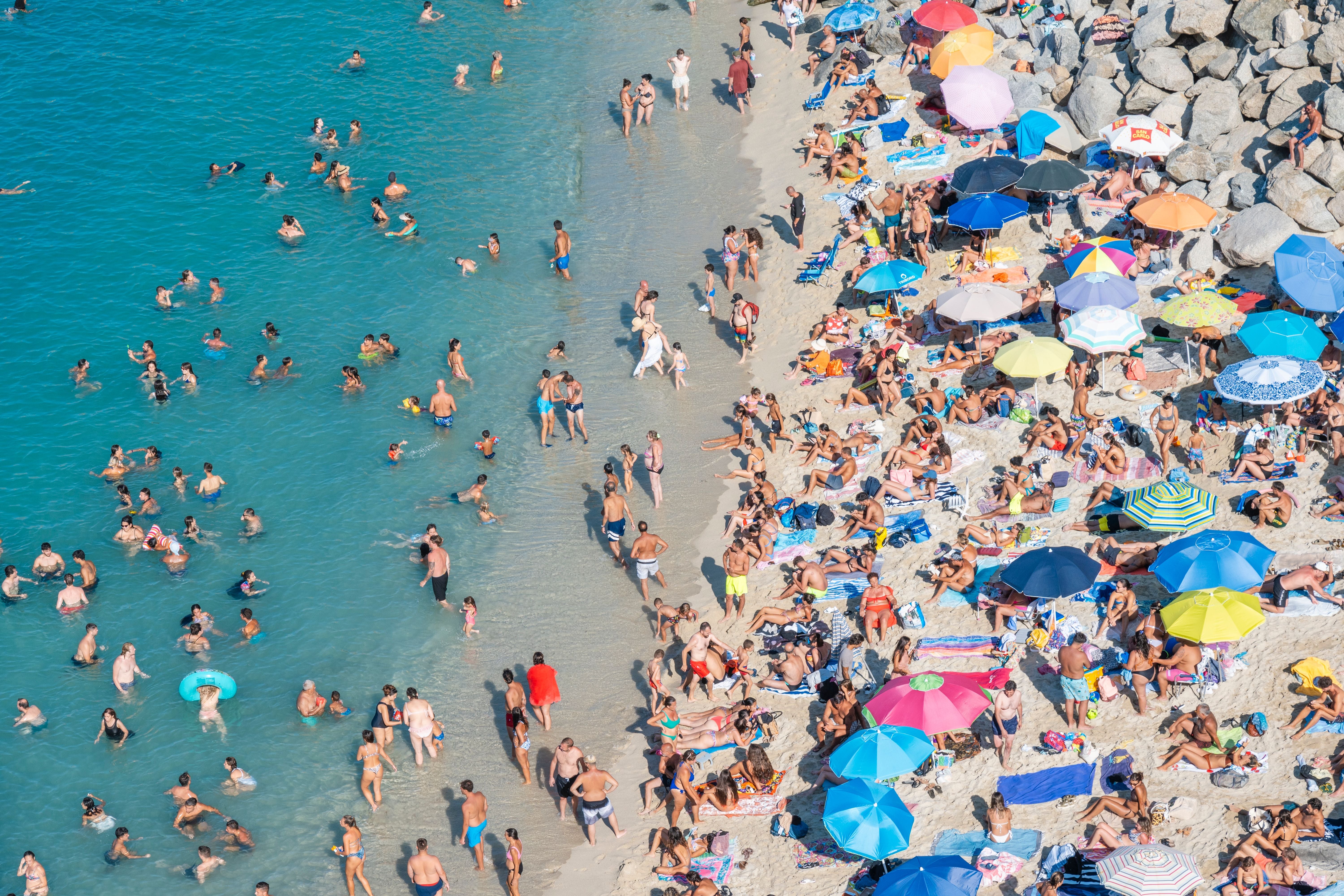 Überfüllter Strand mit vielen Badenden im Wasser und bunten Sonnenschirmen auf dem Sand.