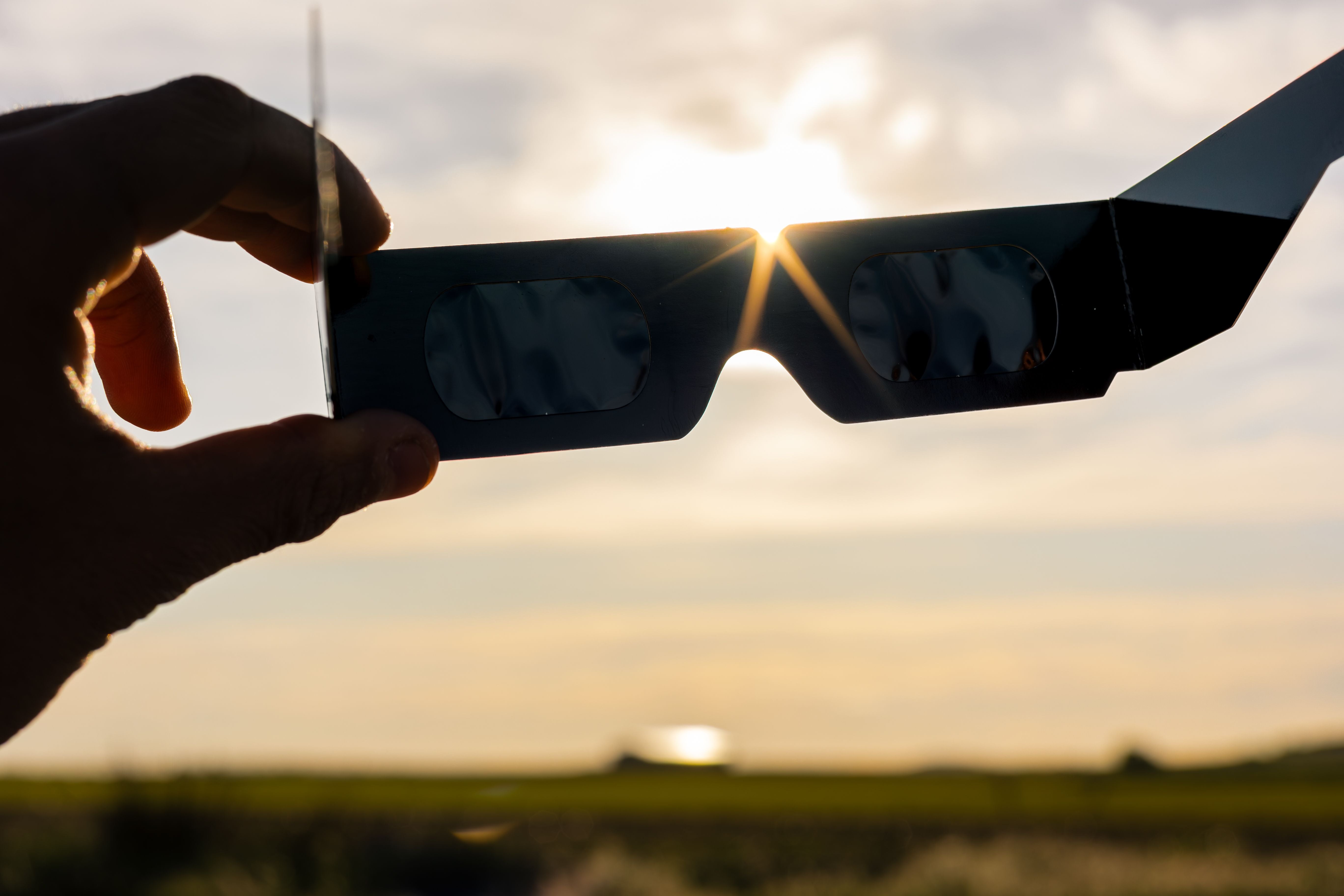 Hand hält Sonnenfinsternisbrille vor die Sonne bei heller Landschaft im Hintergrund.