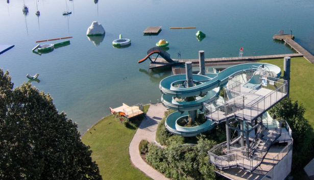 Große Wasserrutsche am See mit schwimmenden Spielgeräten und Steg im Hintergrund.