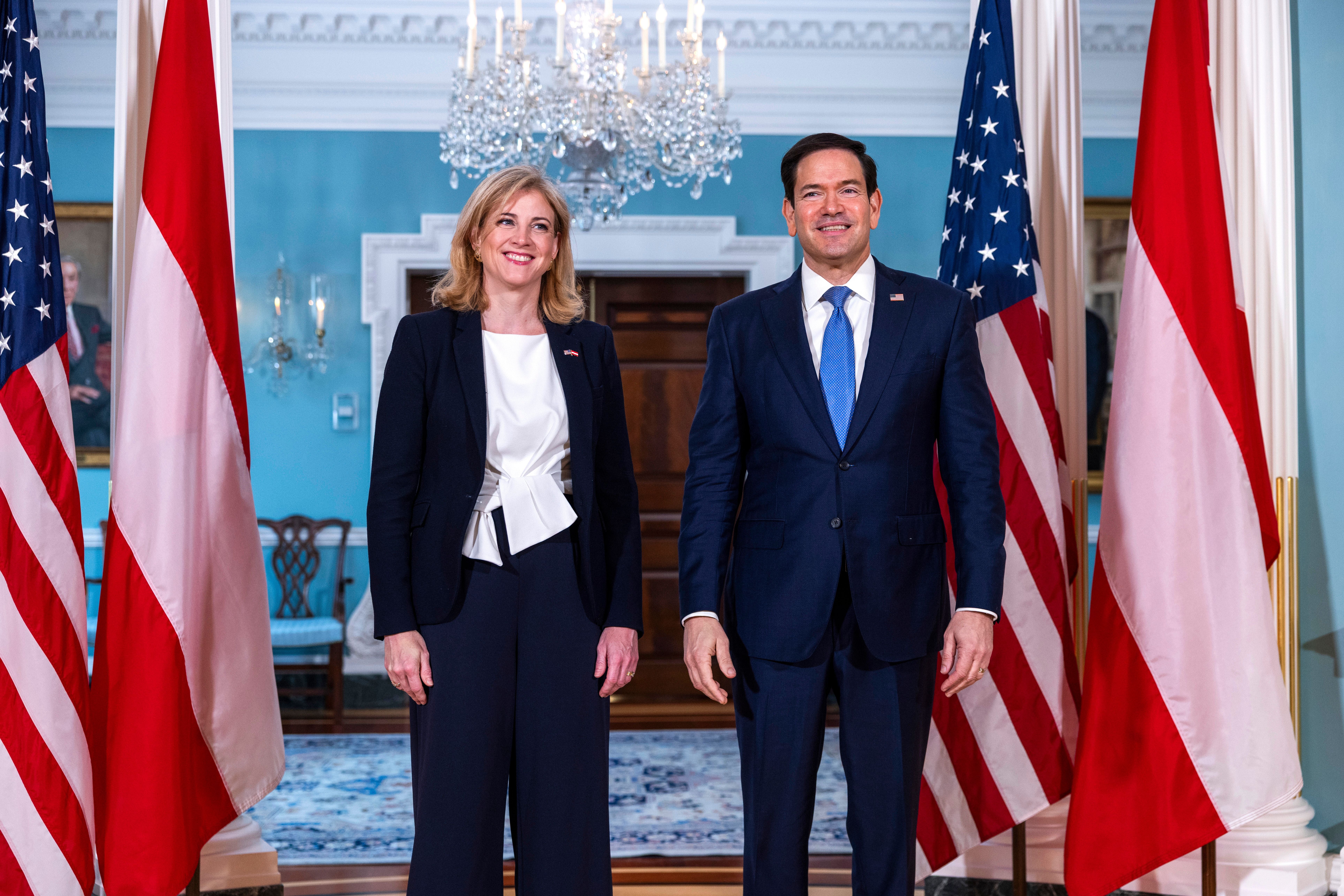 Zwei Personen posieren vor US- und österreichischen Flaggen in einem prunkvollen Raum.