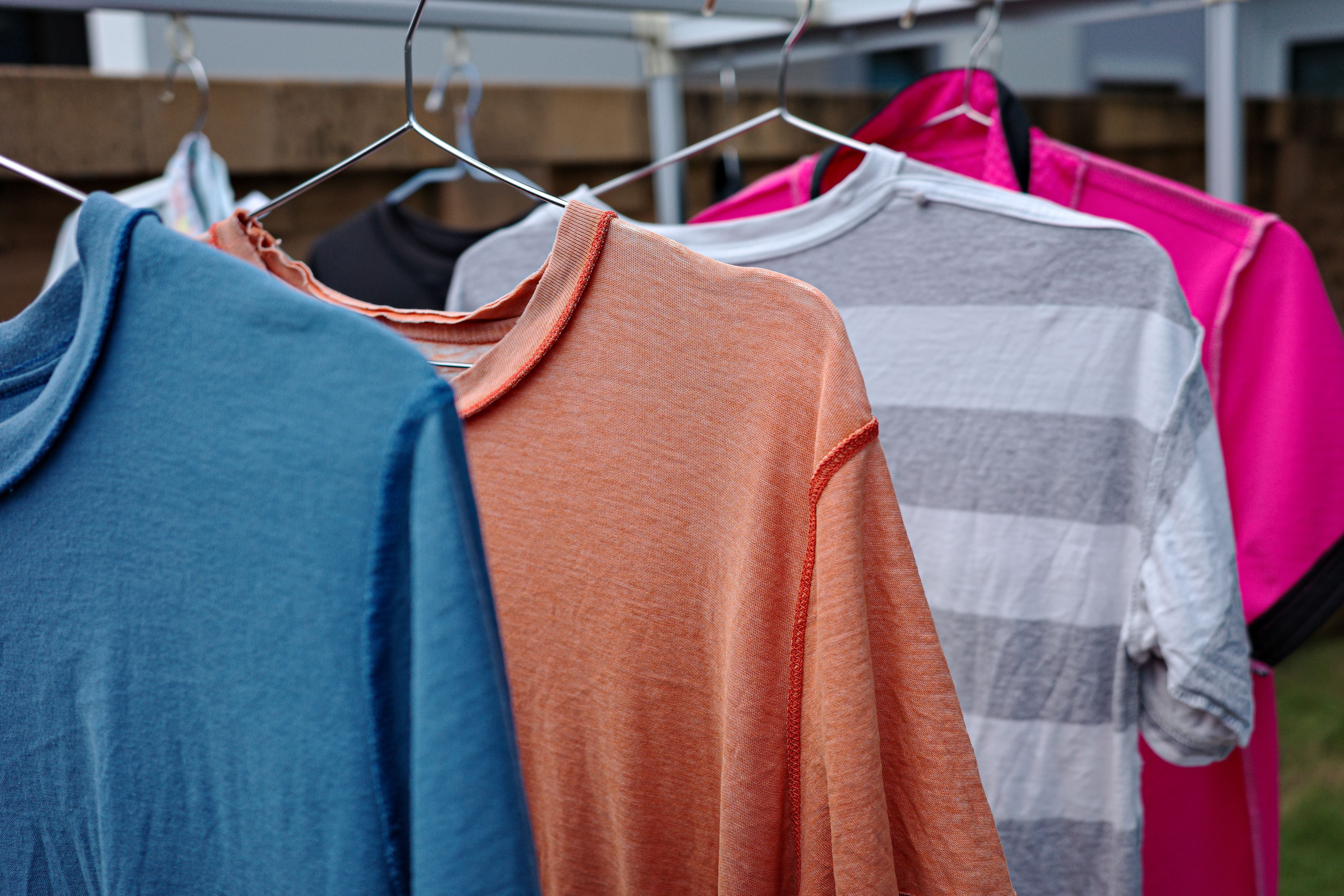 Mehrere bunte T-Shirts hängen auf Kleiderbügeln an einer Wäscheleine im Freien.