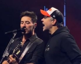 Zwei Männer singen gemeinsam auf einer Bühne, einer spielt Gitarre.