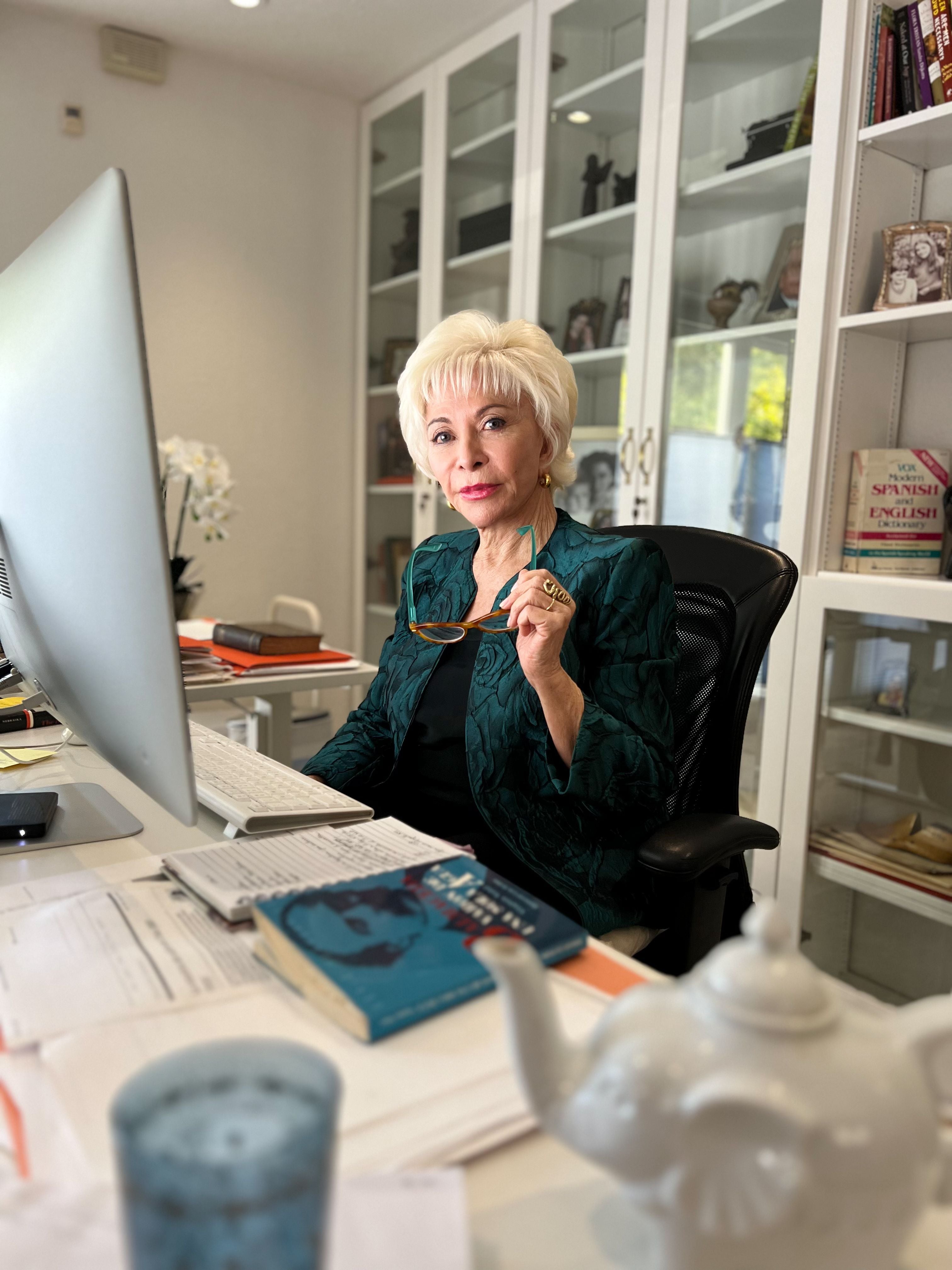Ältere Frau mit blonden Haaren sitzt an einem Schreibtisch mit Computer und Büchern im Homeoffice.