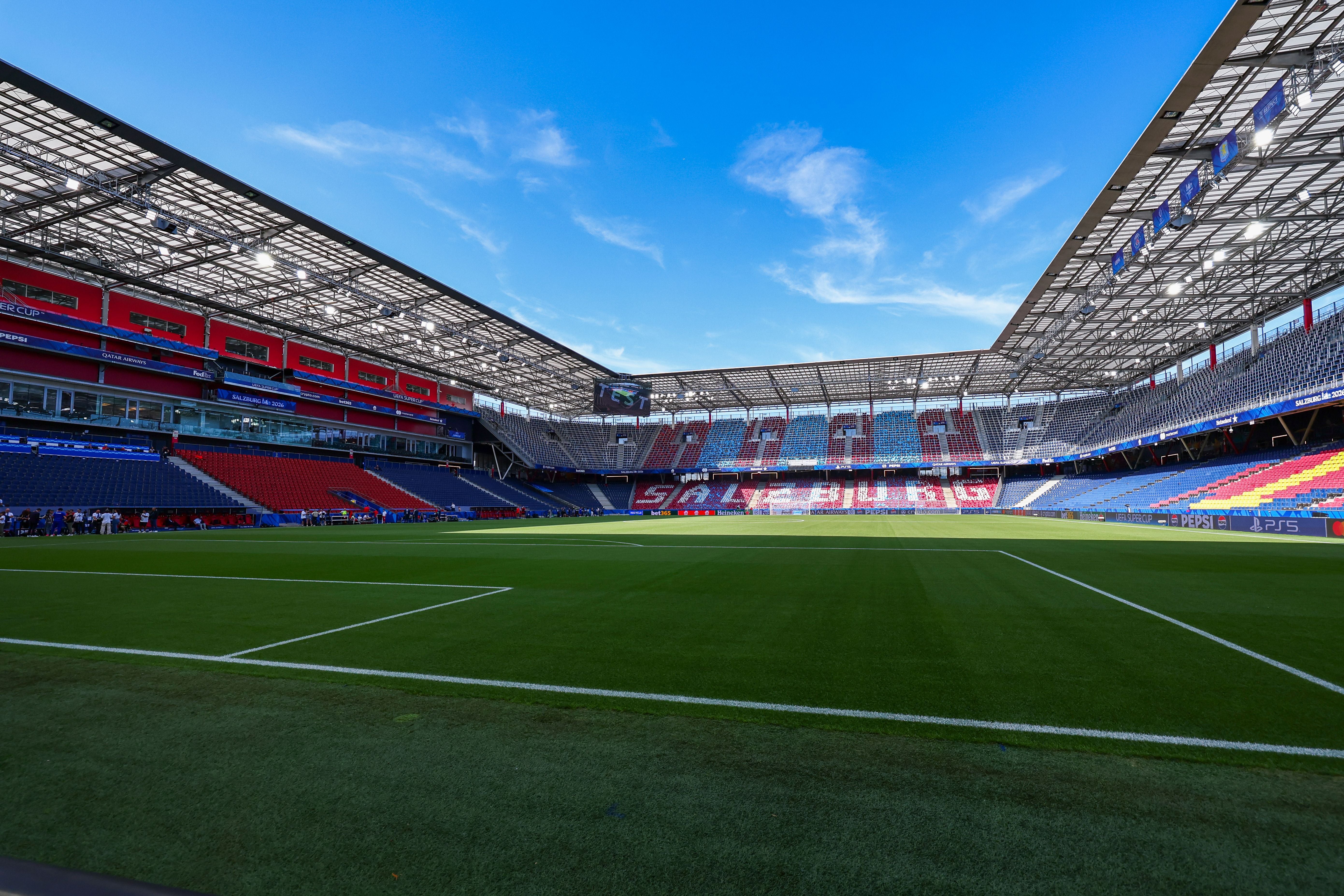 Blick ins leere Salzburg Stadion mit bunten Sitzreihen und grünem Spielfeld unter blauem Himmel.