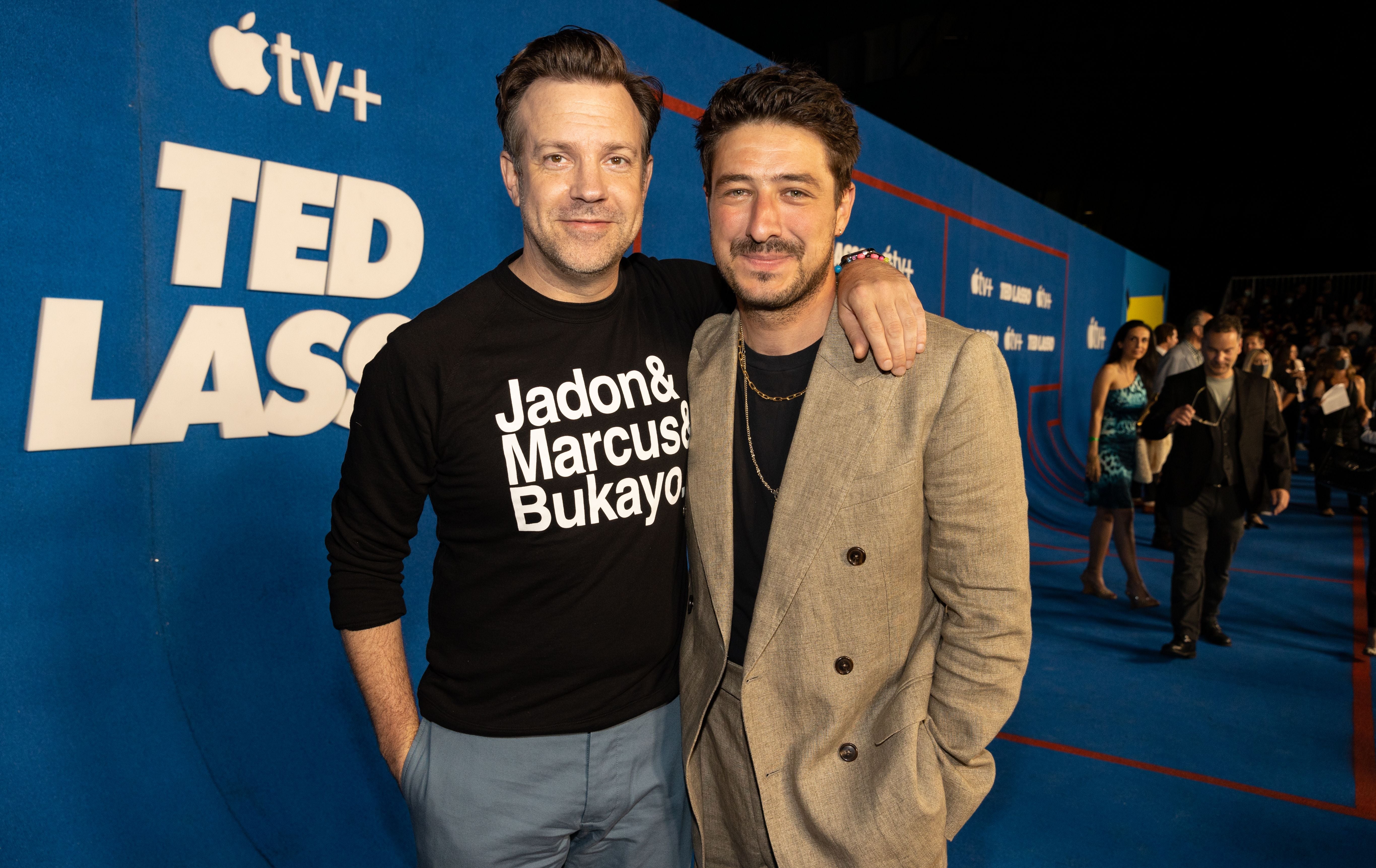 Zwei Männer posieren lächelnd auf dem blauen "Ted Lasso"-Premieren-Teppich.