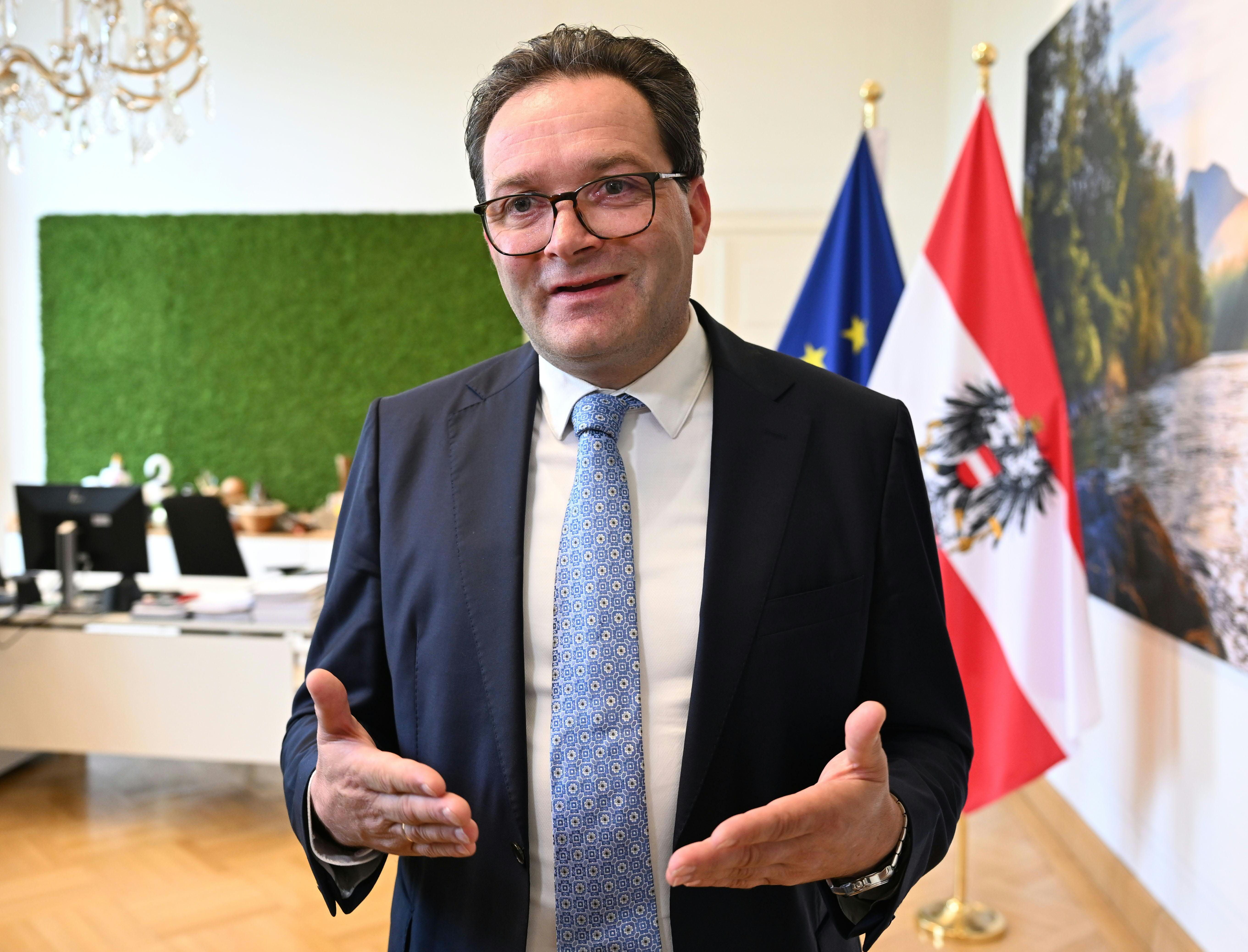 Ein Mann im Anzug spricht in einem Büro vor österreichischer Flagge und EU-Flagge.