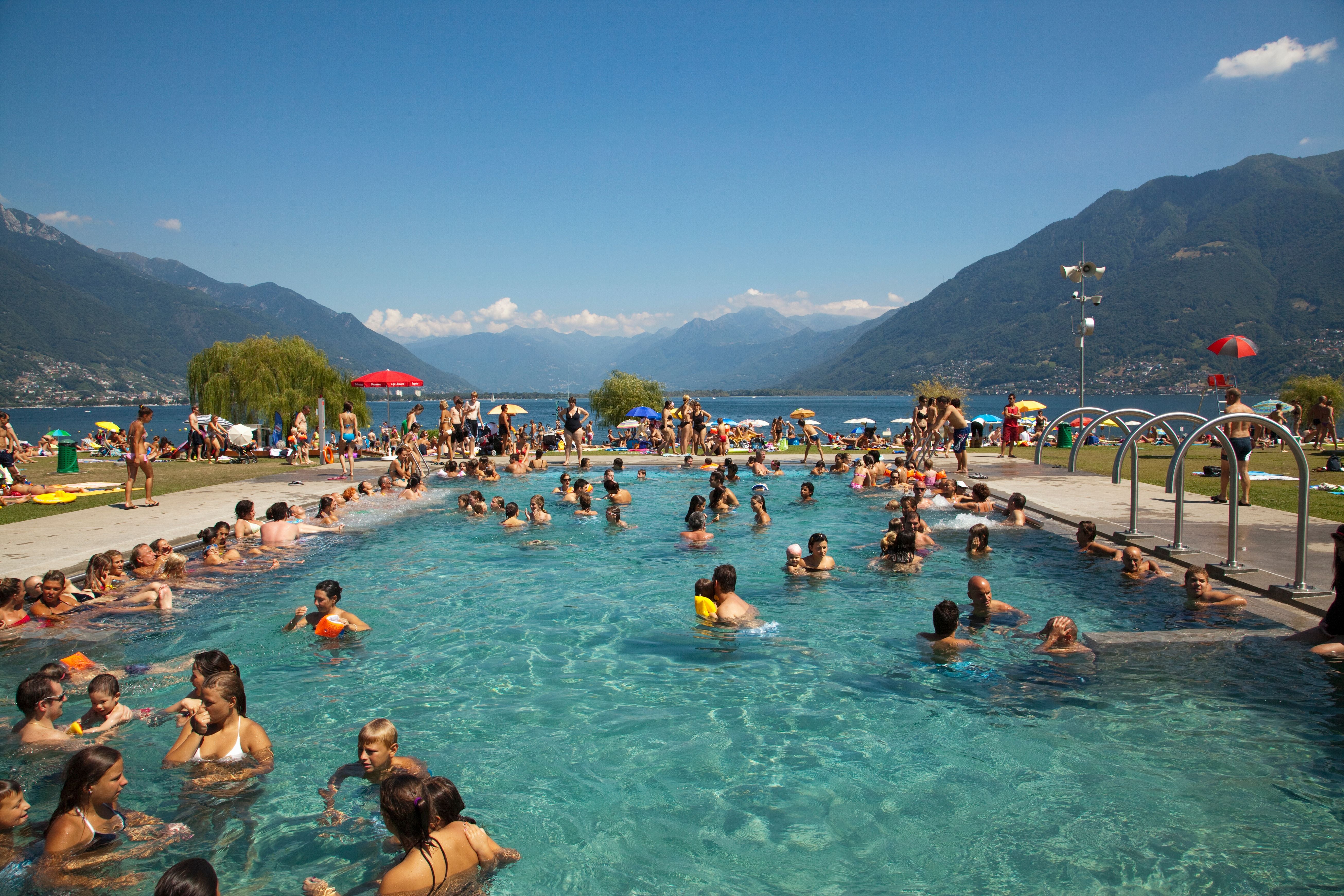 Viele Menschen baden in einem Freibad vor einer Kulisse aus Bergen und einem See bei sonnigem Wetter.