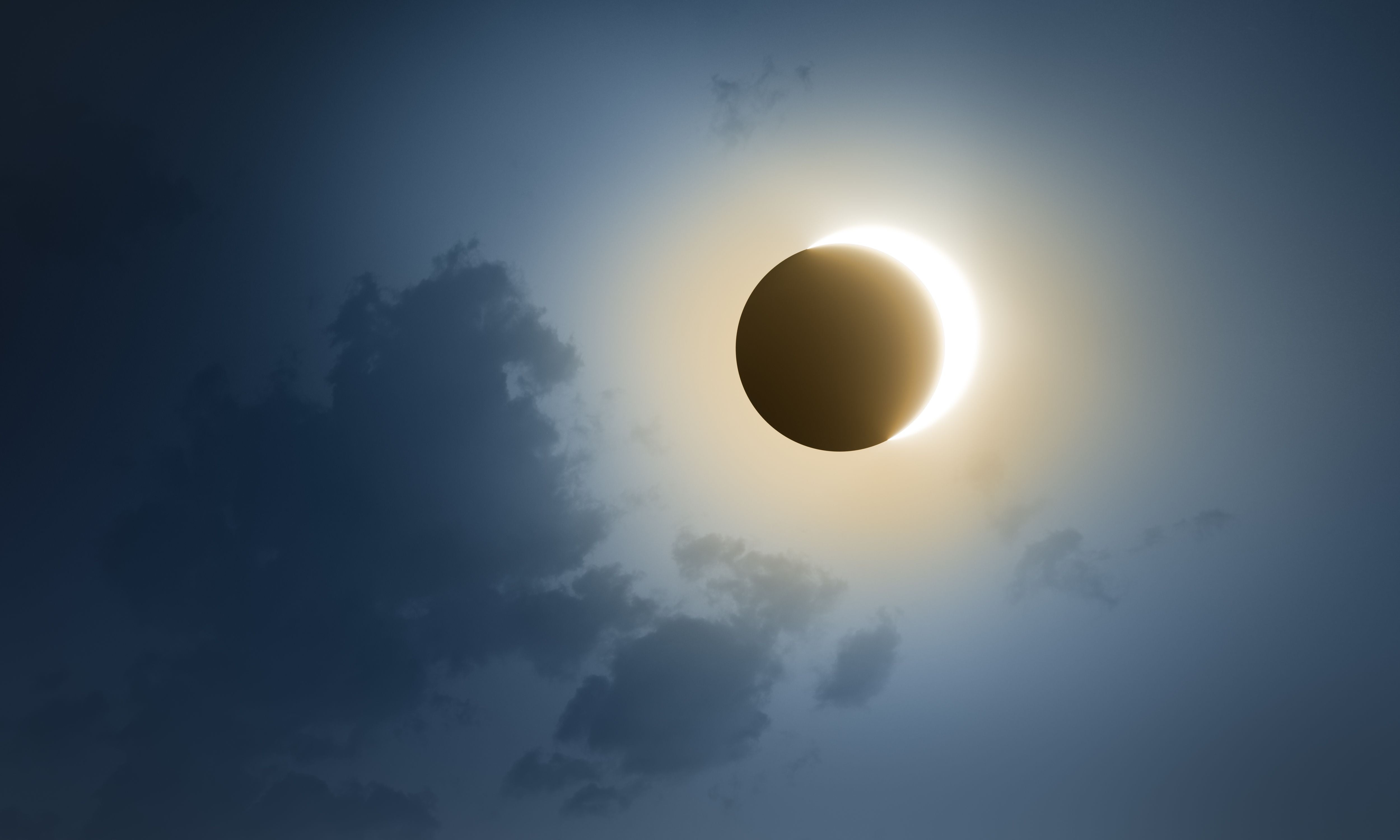 Sonnenfinsternis mit Korona am dunklen Himmel und einigen Wolken im Vordergrund.