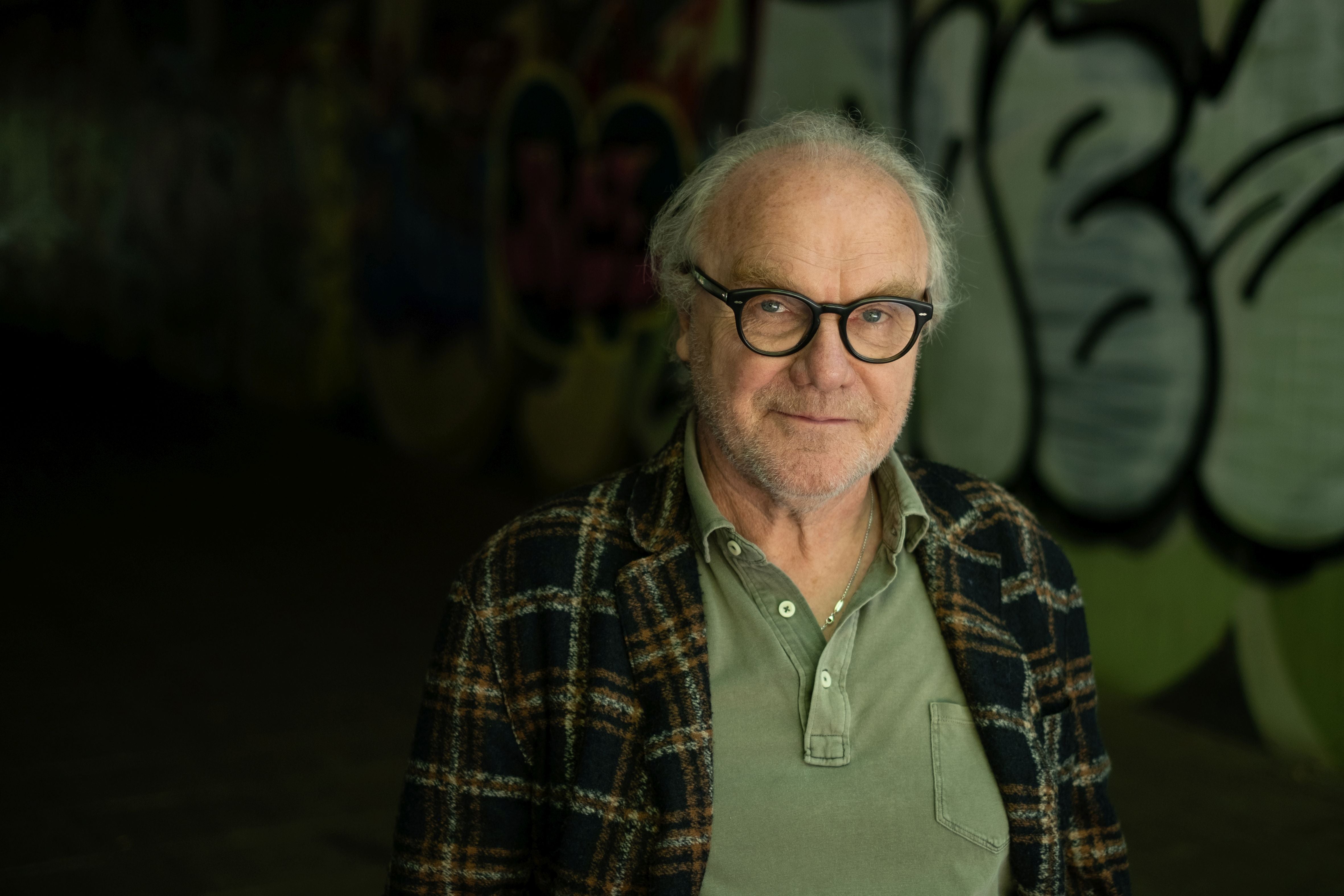 Ein älterer Mann mit Brille steht vor einer Wand mit Graffiti und schaut in die Kamera.
