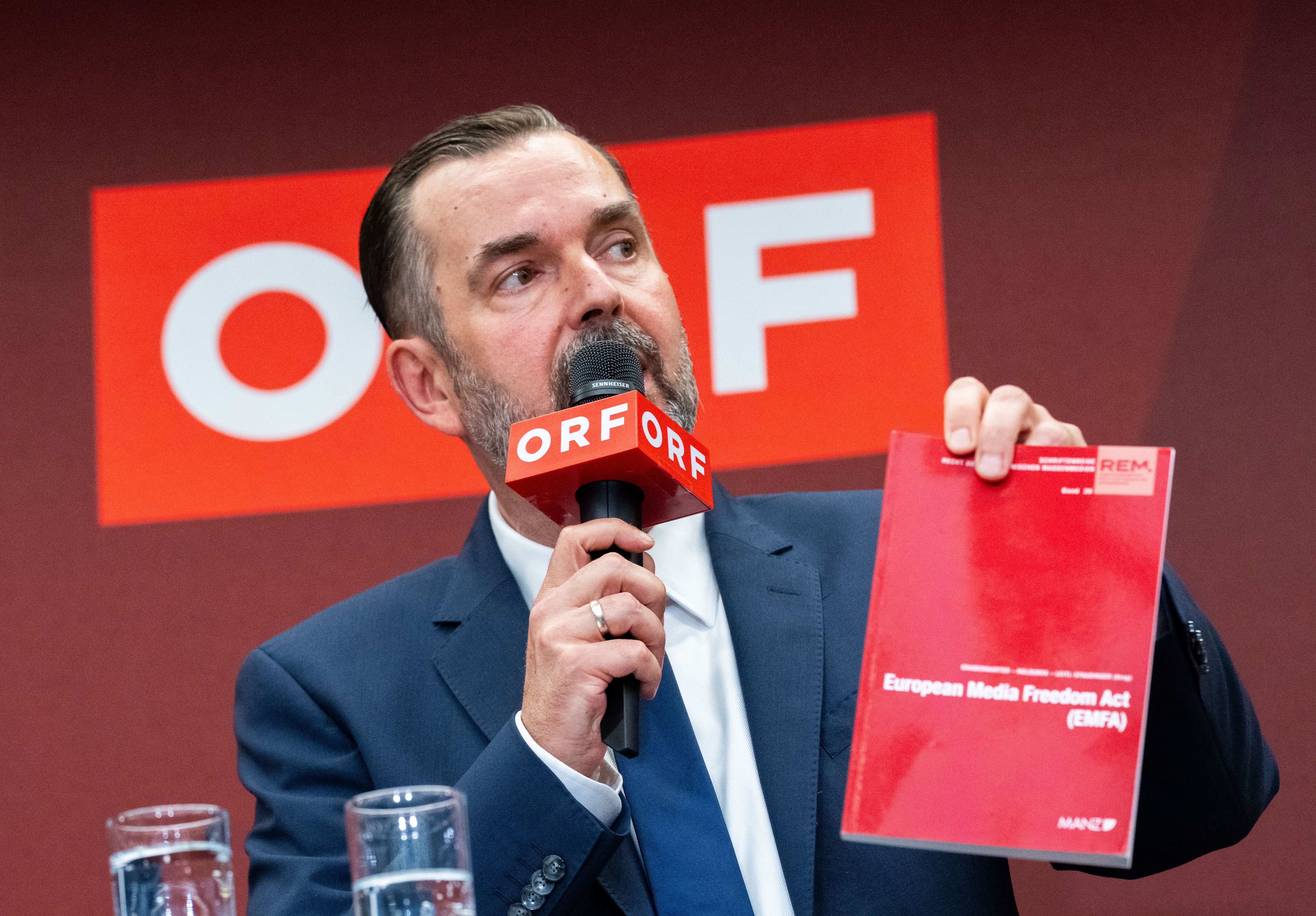 Ein Mann hält ein Mikrofon und ein rotes Buch mit der Aufschrift "European Media Freedom Act".